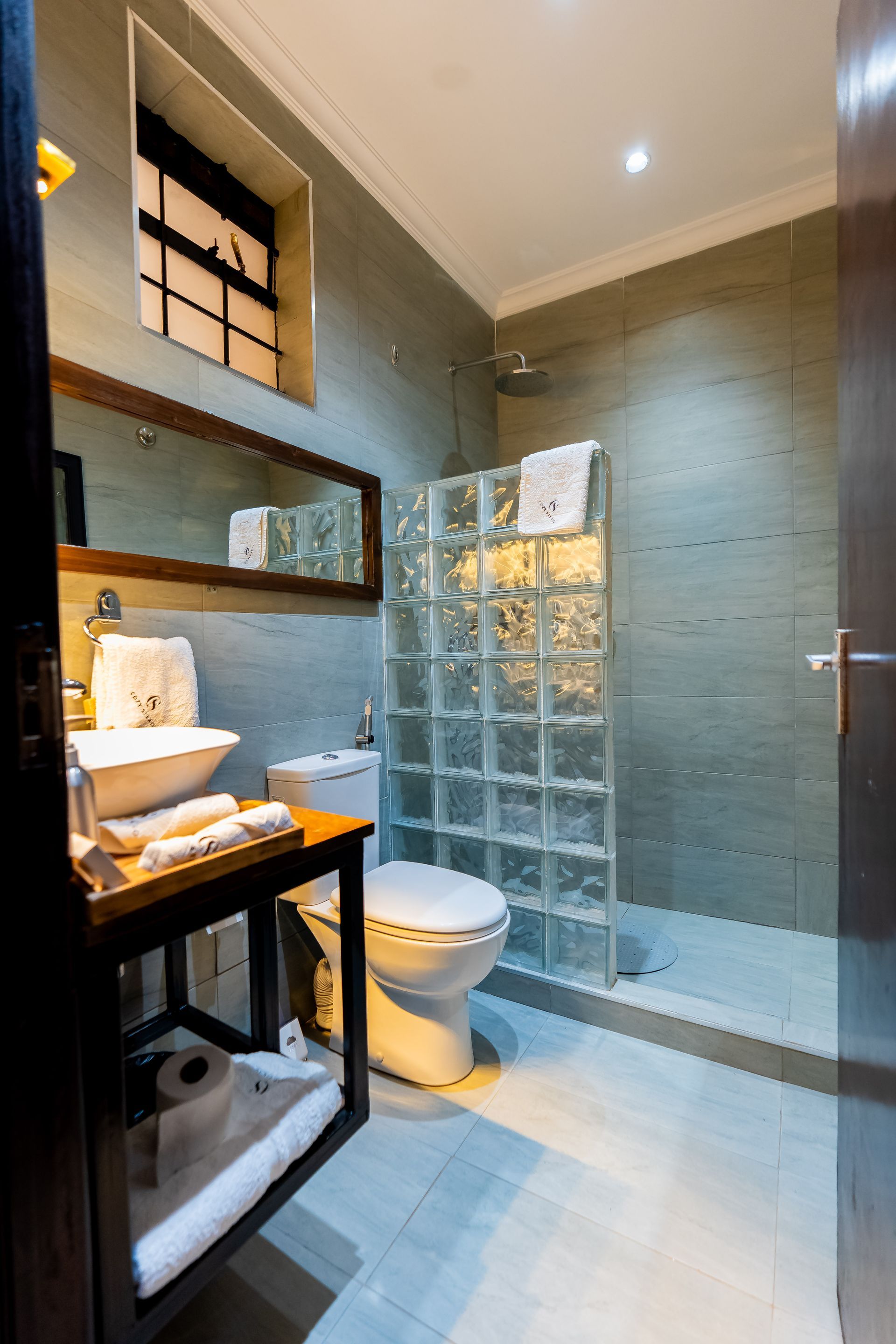 Bathroom with a block shower enclosure, white toilet, sink, and wooden framed mirror. Gray tiled walls and light blue floor.