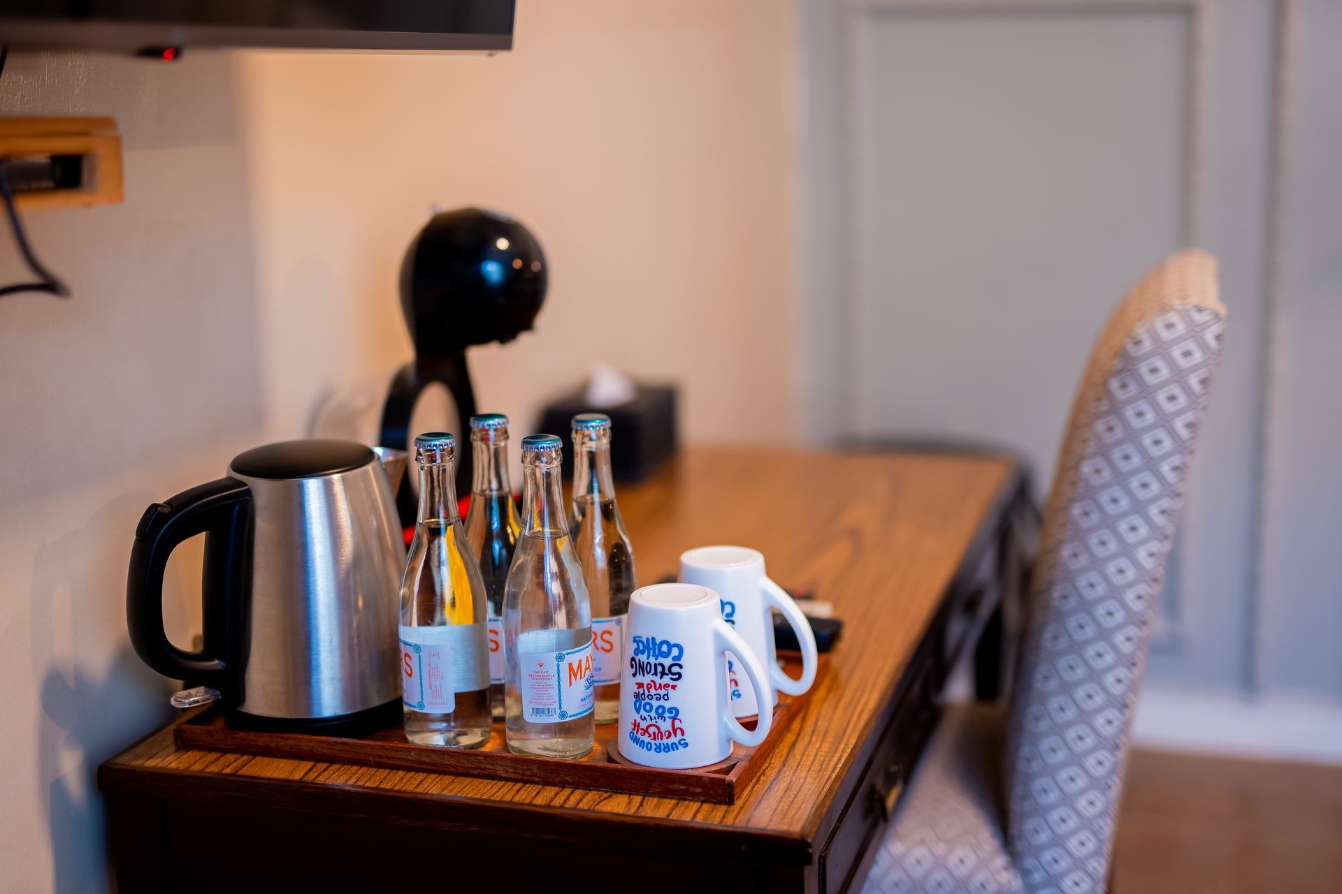 A hotel room desk with a water kettle, bottles of water, coffee maker, mugs, and a patterned chair.
