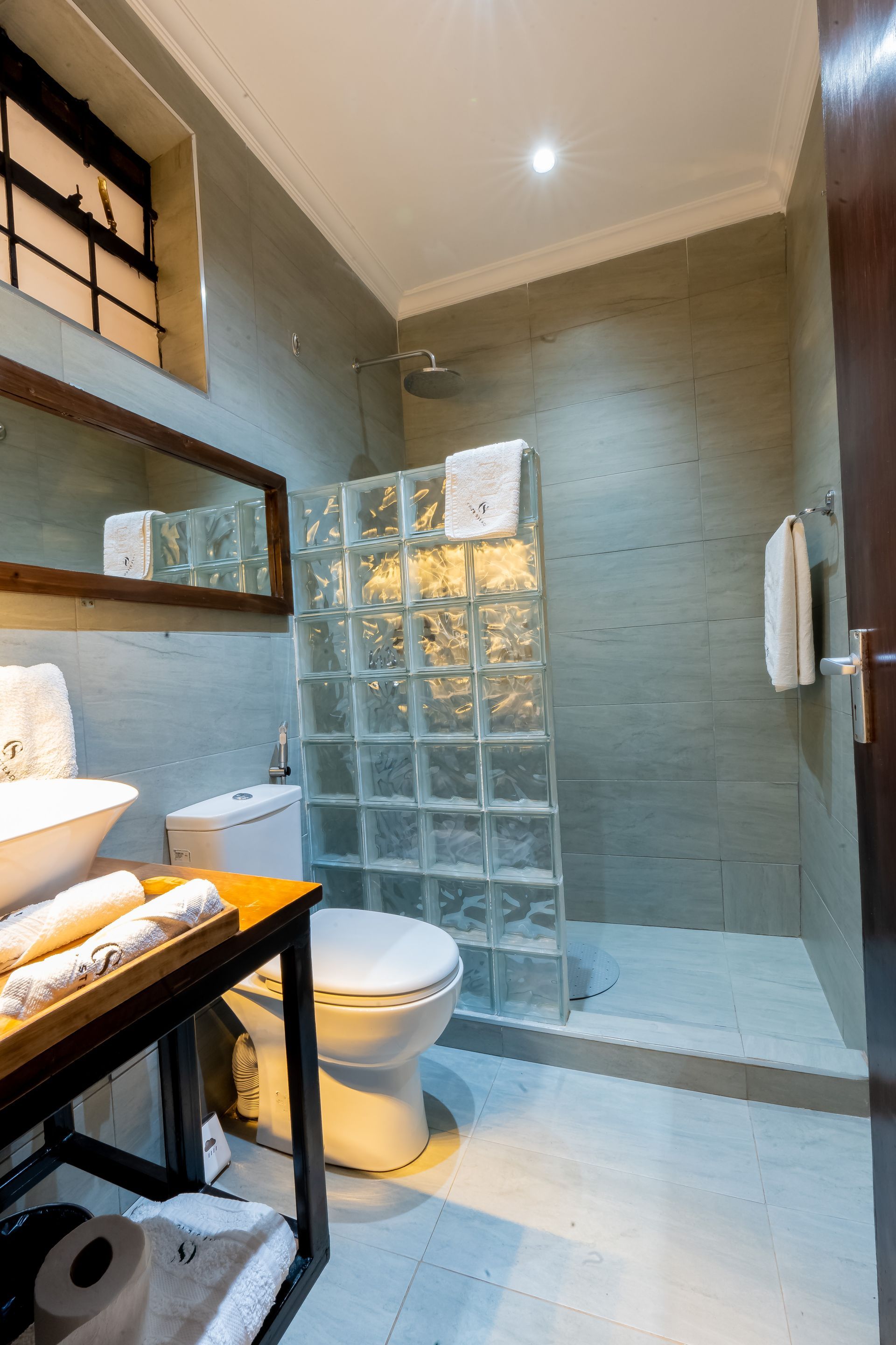 bathroom with a toilet, shower and vanity. A brick wall separates the shower area. The walls are gray with white towels.