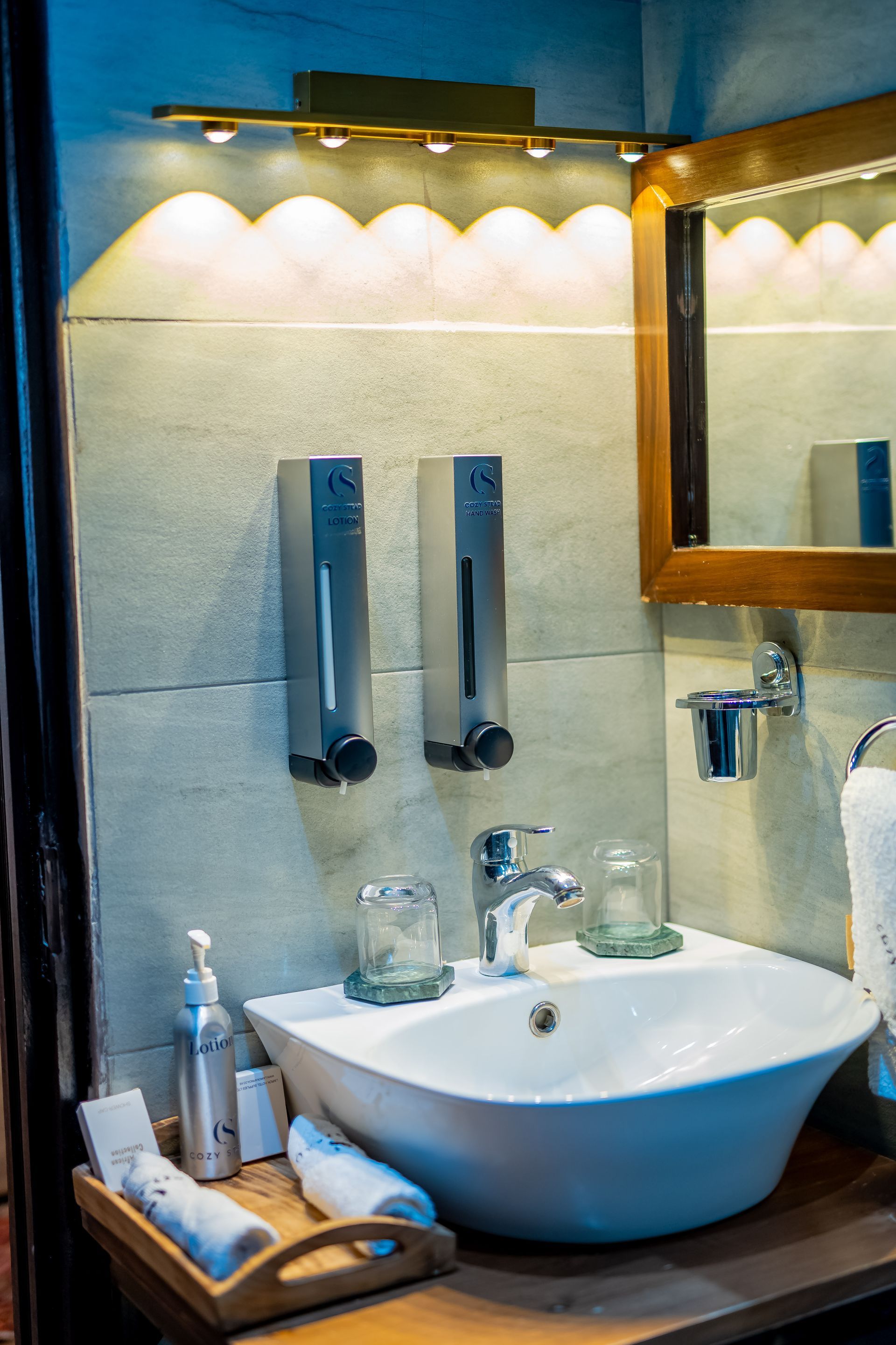 A bathroom sink with soap dispensers, mirror, and toiletries. Soft lighting and a wooden shelf create a spa-like atmosphere.