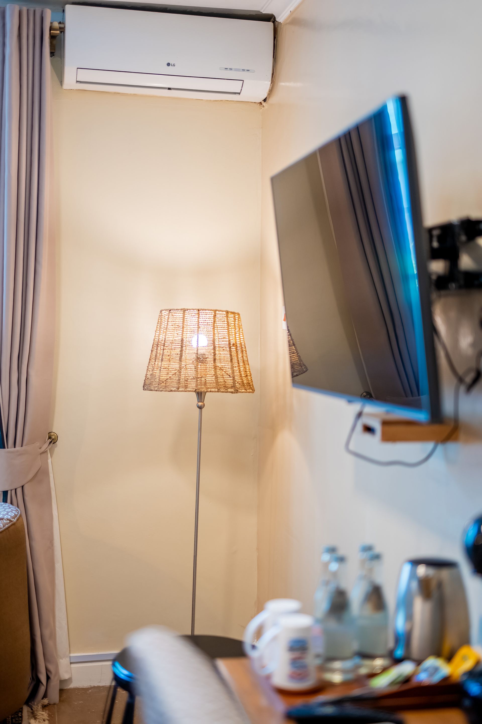 A floor lamp with a textured shade illuminates a corner of a room with a wall-mounted TV and mini bar.