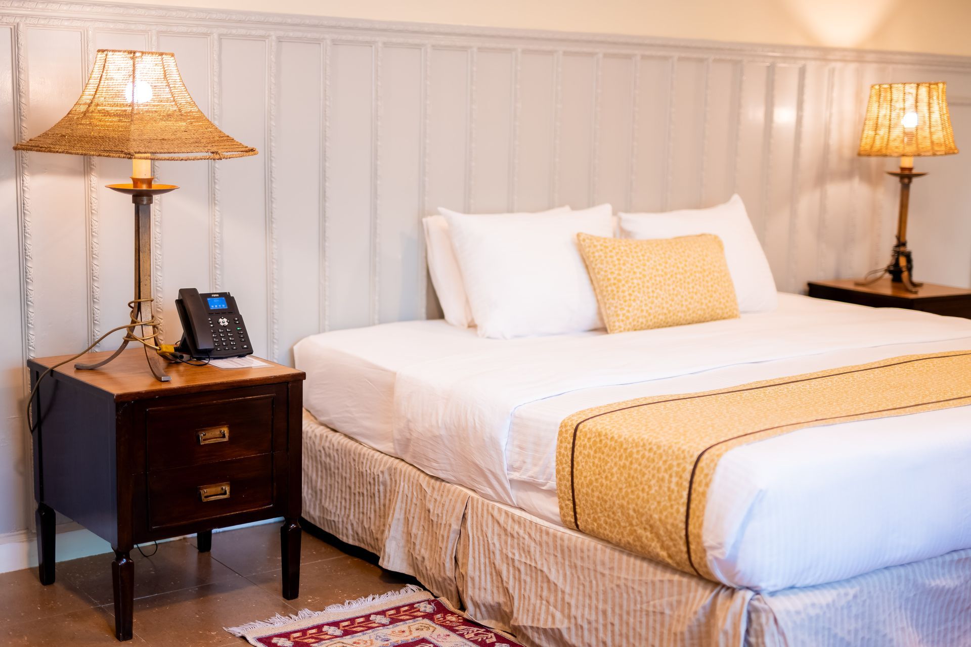 Cozy hotel bedroom with a bed, lamps, and a nightstand. Yellow and white bedding, wood paneling, and a small rug are visible.