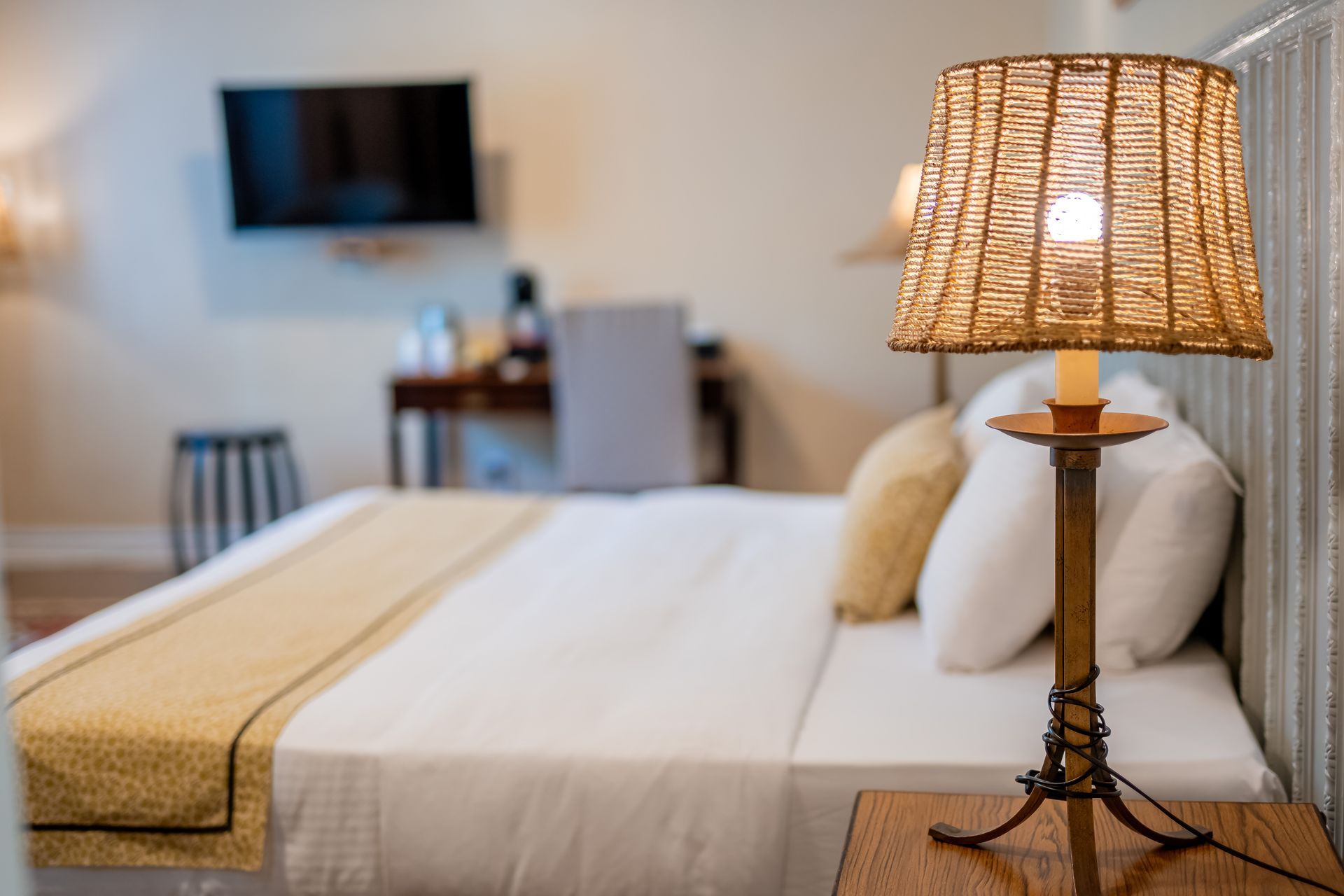 Bedroom with a bed, lamp on a nightstand and a TV on the wall. The bed has white linens and gold accents, with a wicker lamp.