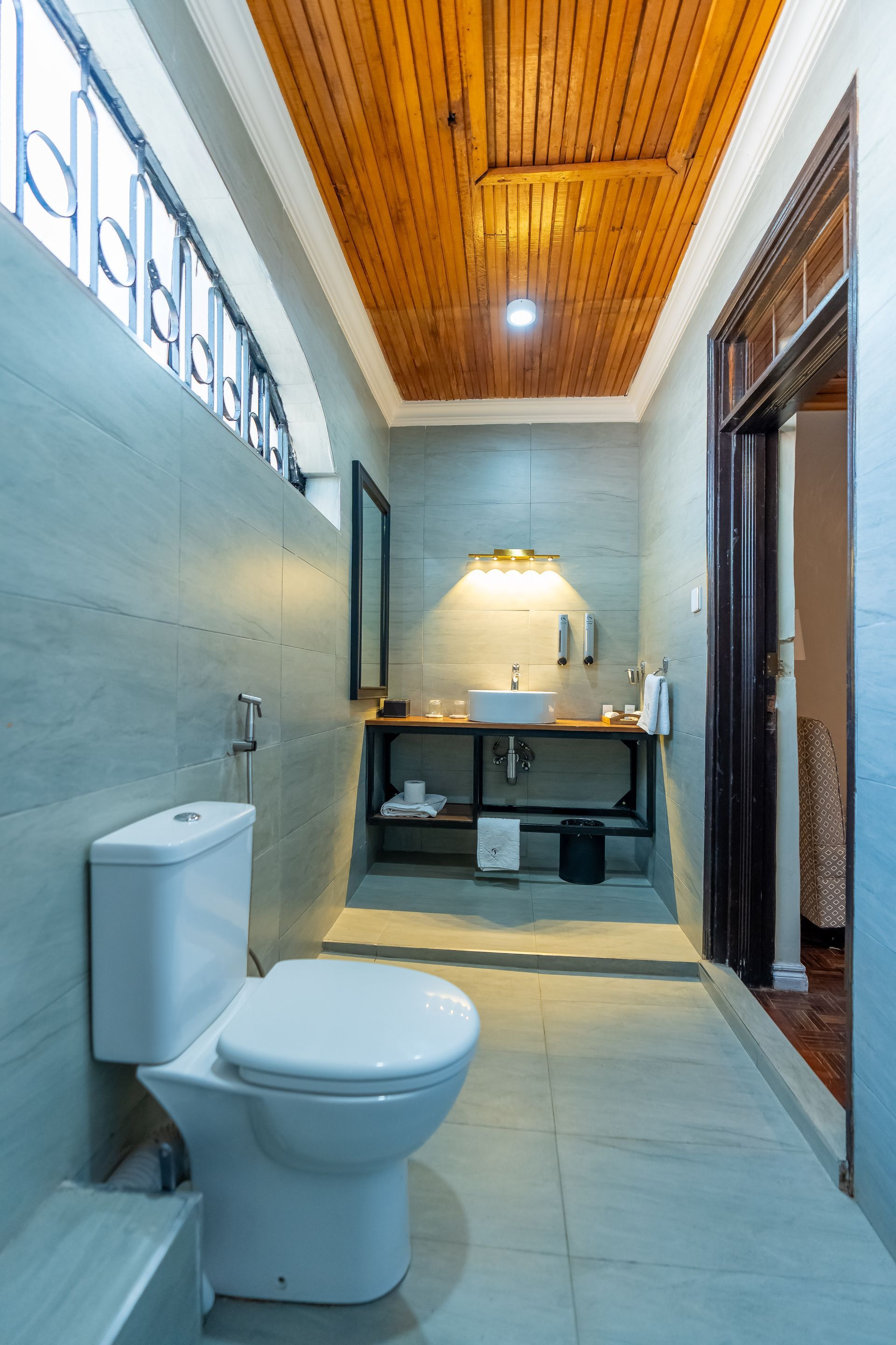 A bathroom featuring a toilet, sink, and a wooden ceiling. Light-colored walls and a window with a decorative railing.