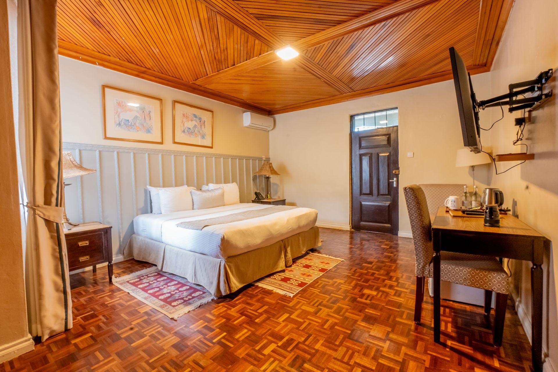 hotel room with a king-sized bed, wooden floors, and a desk area. The room has a wood-paneled ceiling and is brightly lit.