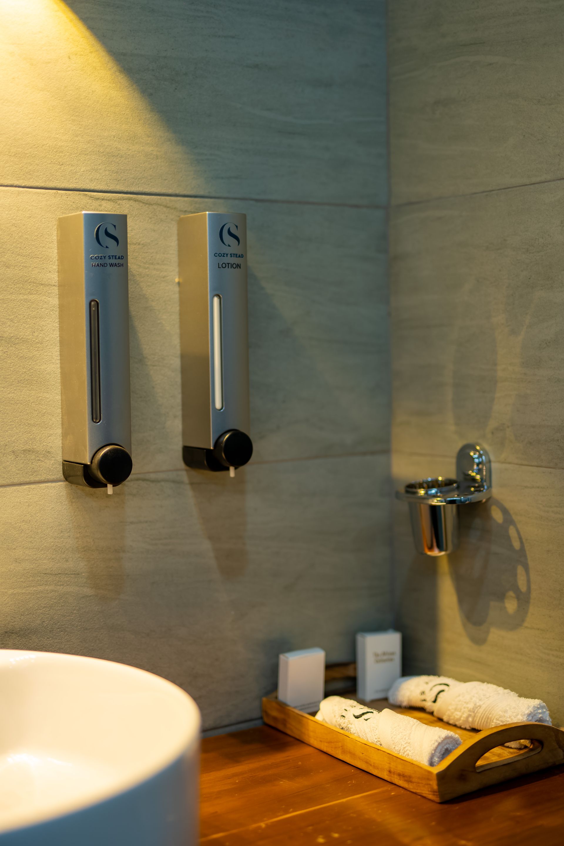 Two soap dispensers and a toothbrush holder are mounted on a  wall above a countertop.A tray holds towels and toiletries.