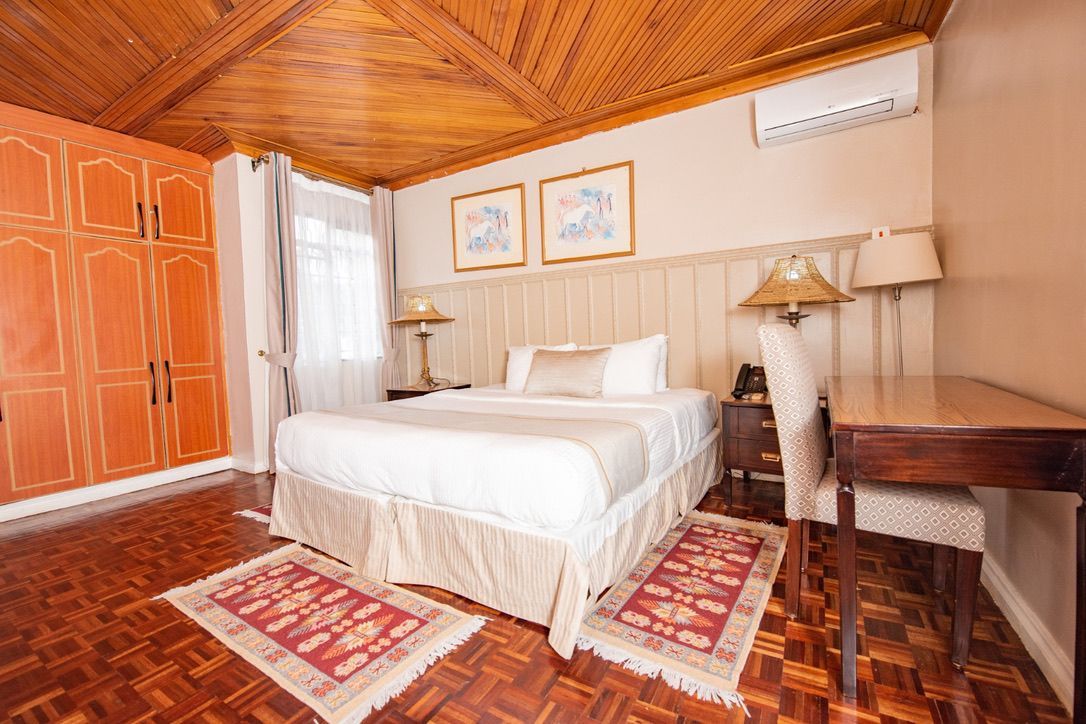 A hotel bedroom featuring a bed with a white comforter, wooden desk, and wardrobe, and patterned rugs on a wooden floor.