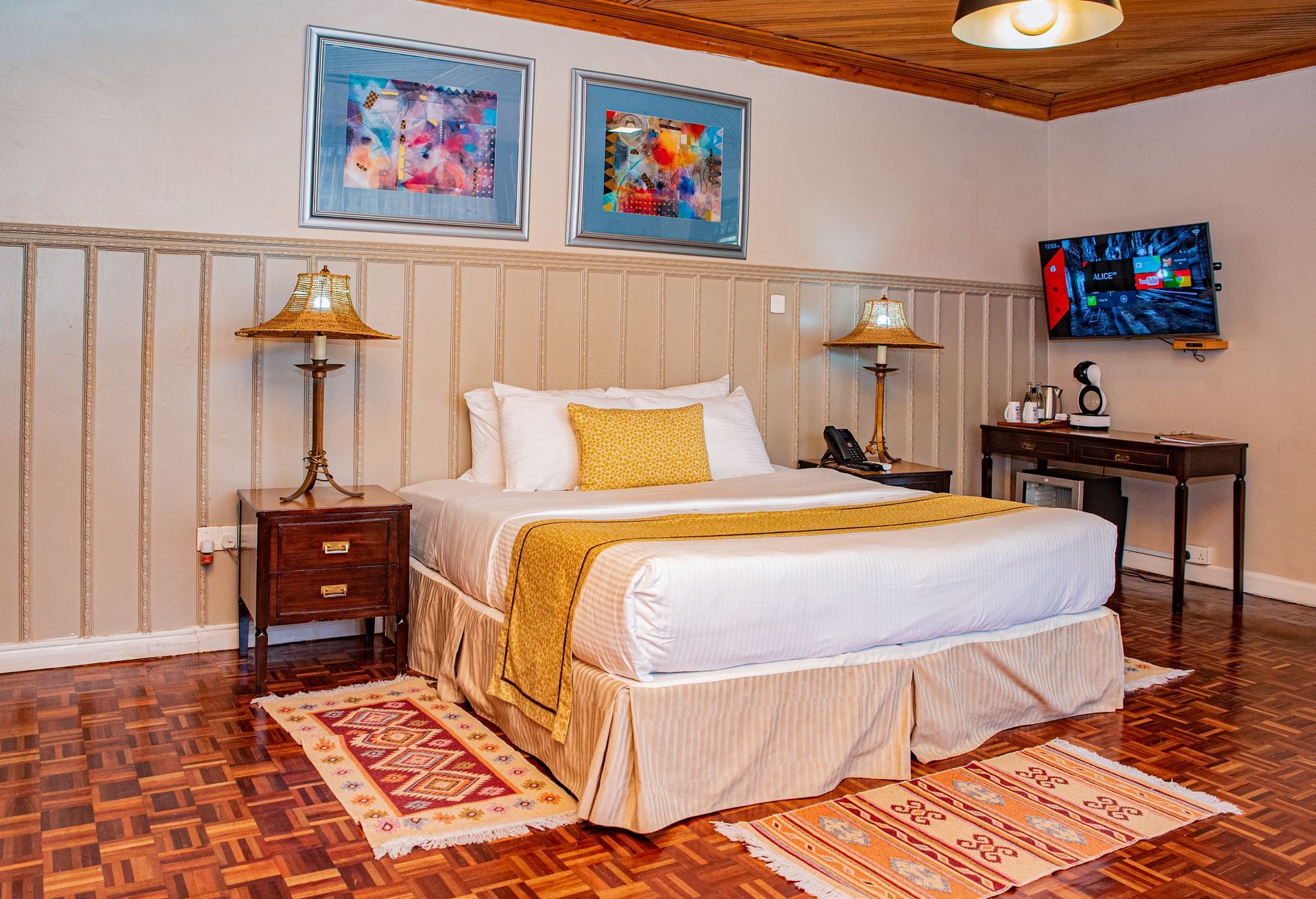 A hotel bedroom with a bed, lamps, artwork, and a television. The room has patterned wooden floors and a decorative ceiling.