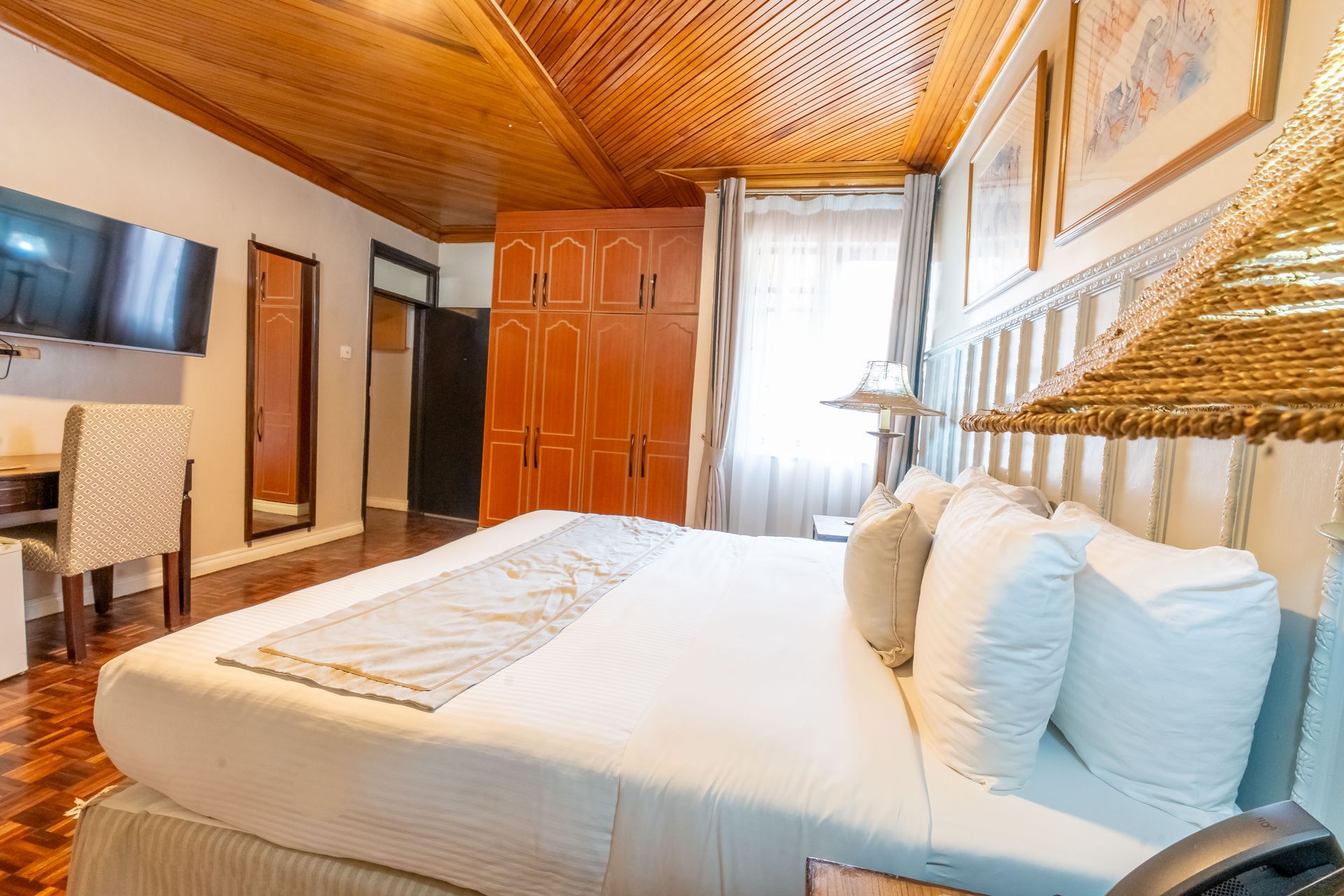 Cozy hotel room with a large bed, wooden ceiling, and warm lighting. Includes a TV, desk, and tall wooden wardrobe.