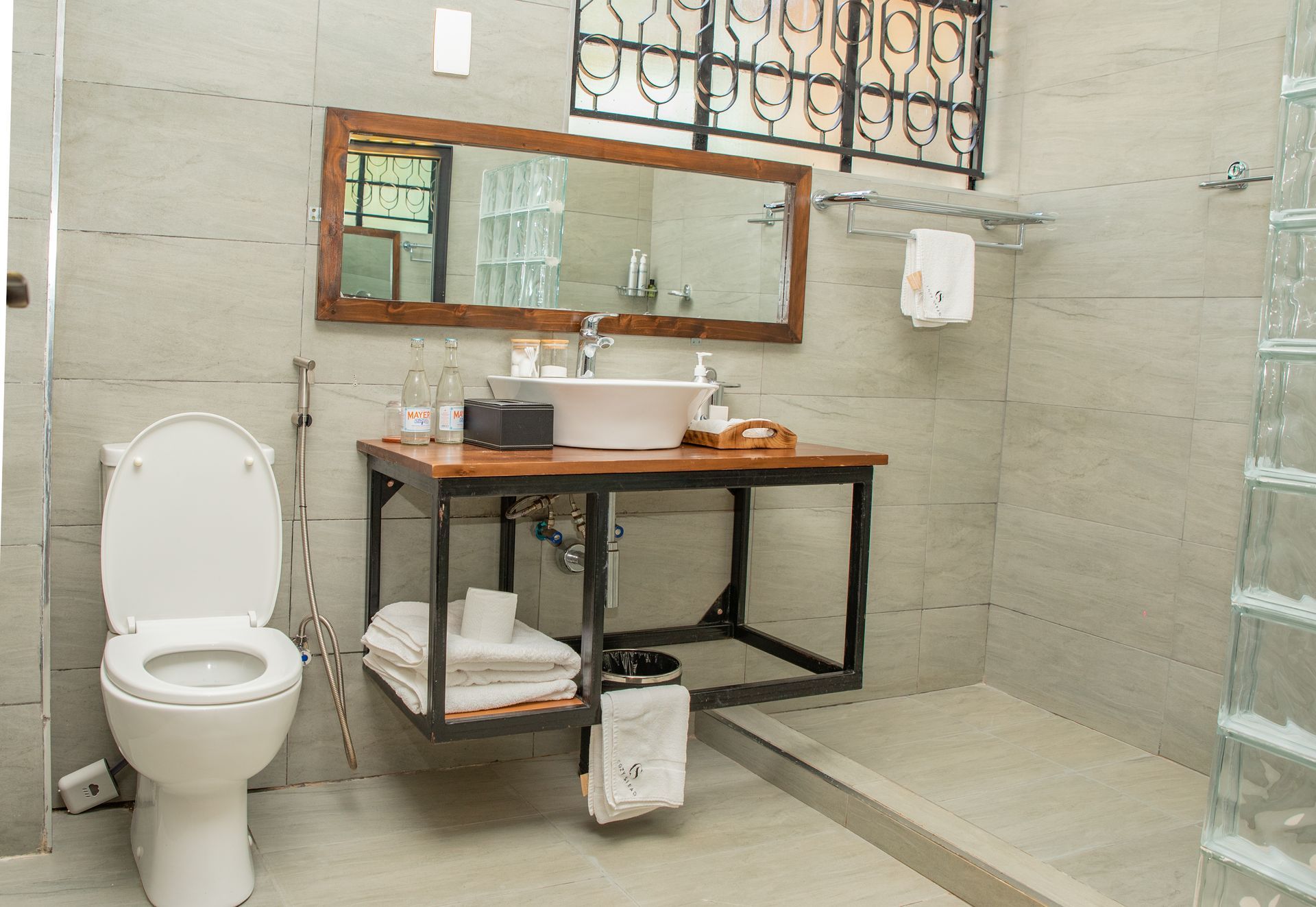 Bathroom with a toilet, sink on a wooden table, and glass block shower. The walls are light grey, and there is a window.