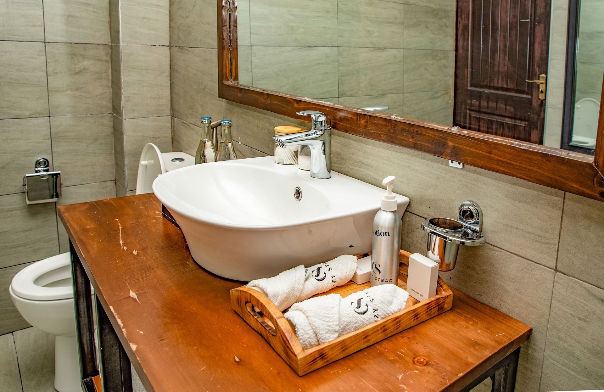 Bathroom with white vessel sink on a wooden vanity, towels in a wooden tray, toilet on the left, and a mirror above.
