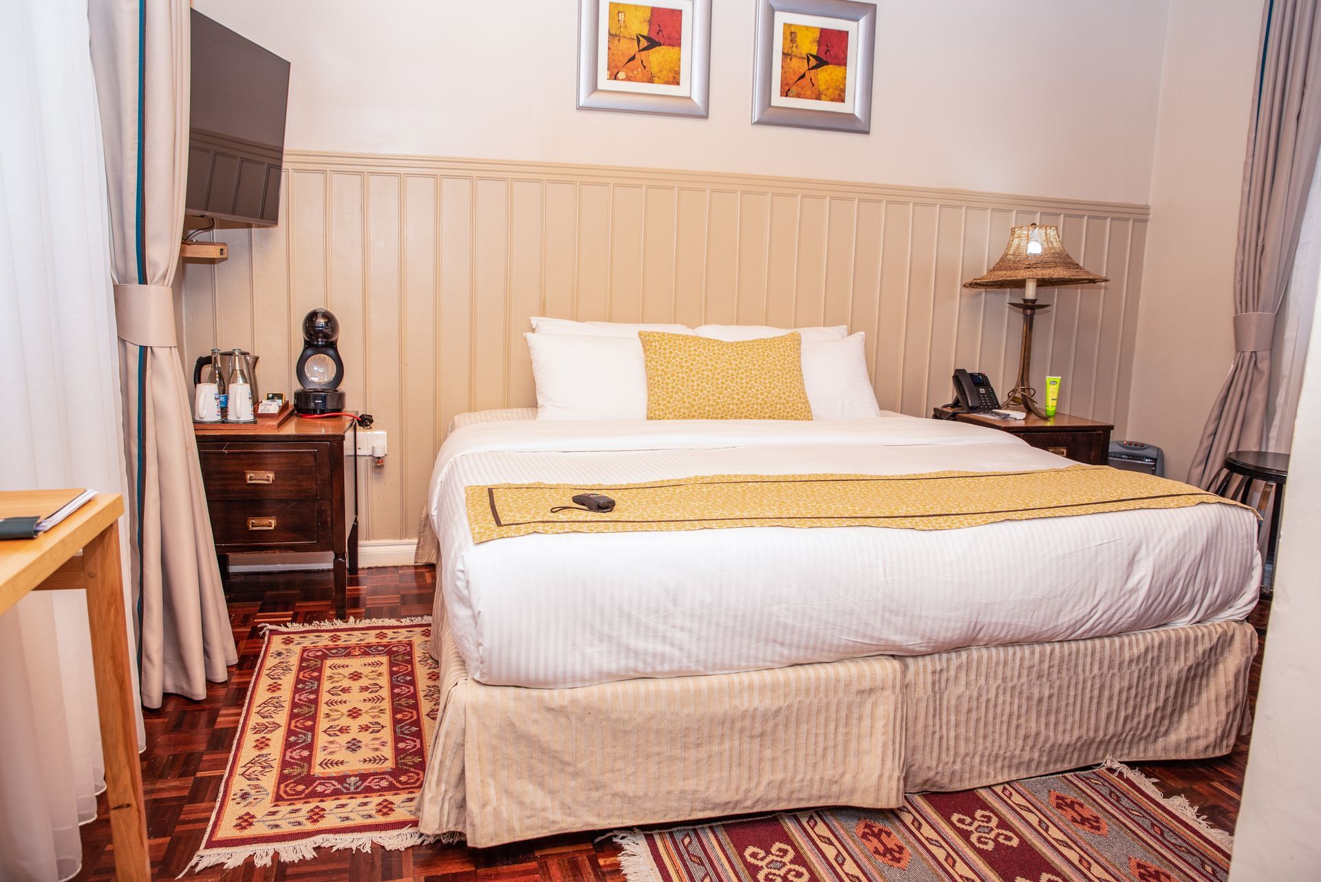 A bedroom with a bed, a nightstand with a lamp, and a TV mounted on the wall. There are also patterned rugs on the floor.