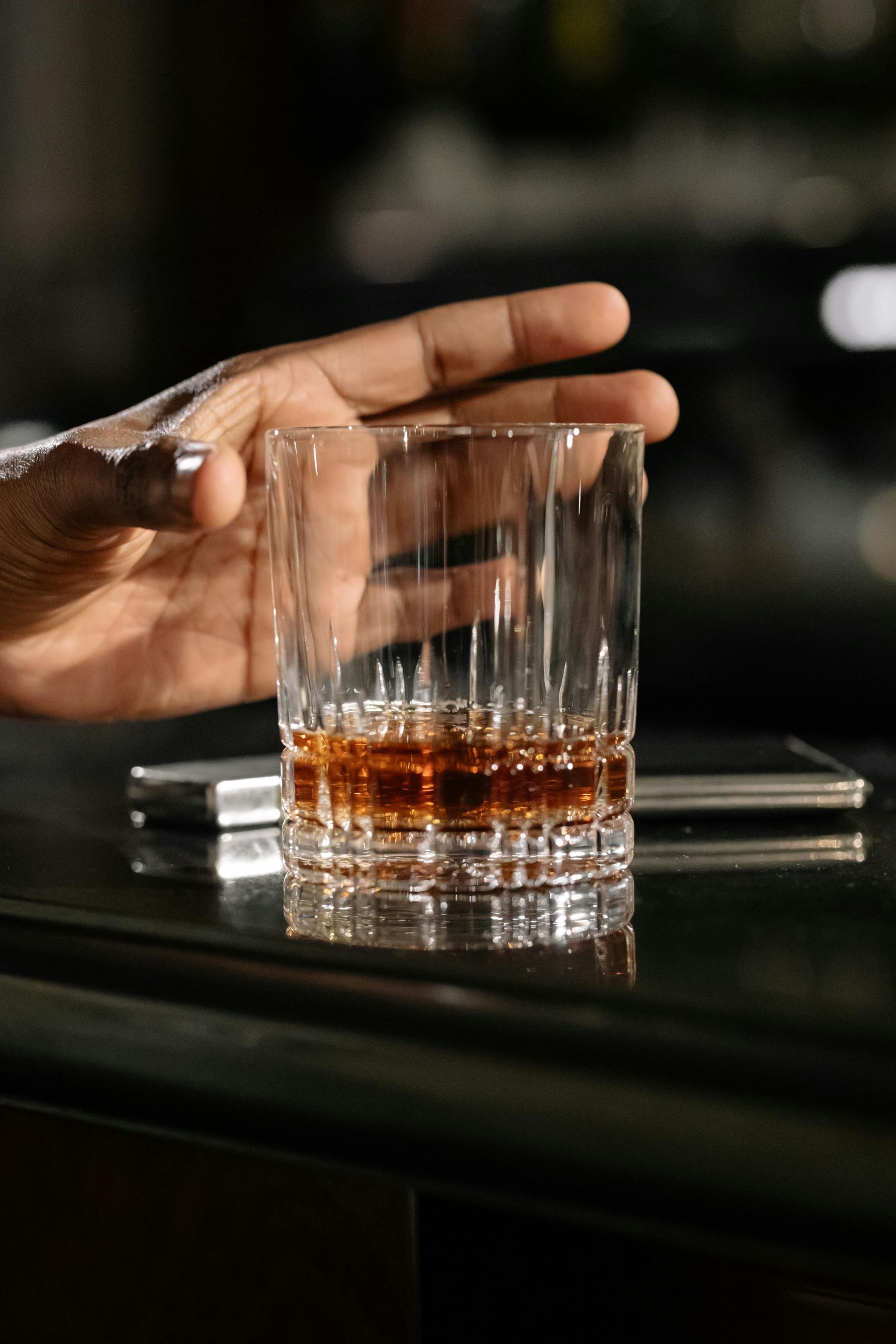 Hand reaching for a rocks glass filled with amber-colored liquor on a bar. A cell phone sits nearby.