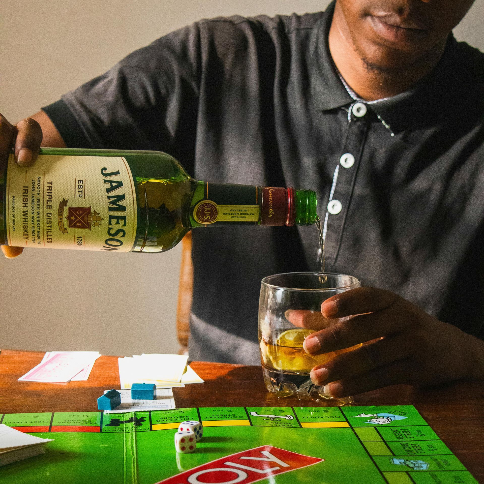 Man pouring Jameson whiskey into a glass while playing Monopoly. A game board, dice, and cards are on the table.