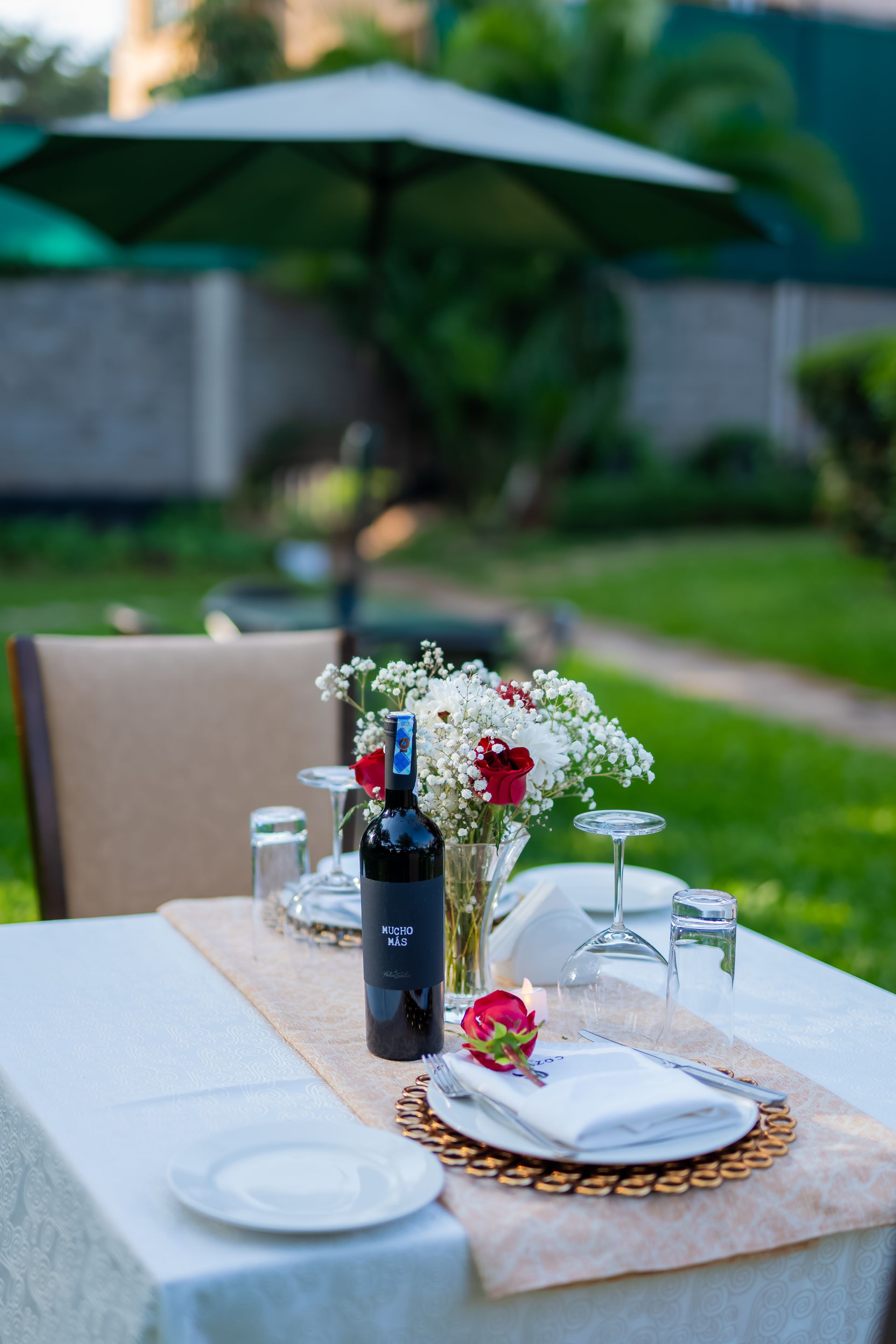 Outdoor table set for a romantic meal: white table, wine bottle, flowers and a napkin with a rose and umbrella in background.