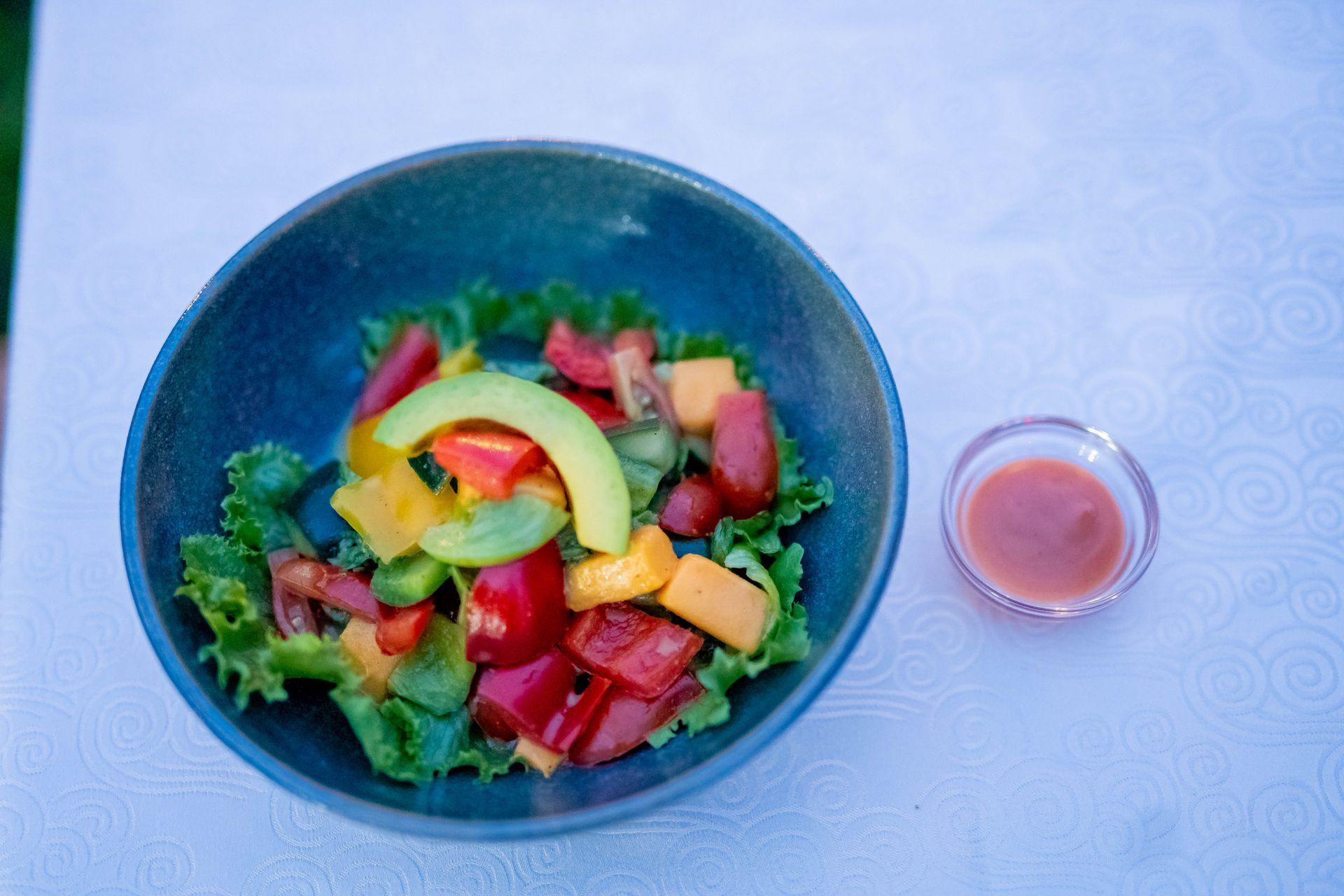 Salad in a bowl with a small dish of dressing next to it on a table. Ingredients include tomatoes, avocado, and peppers.