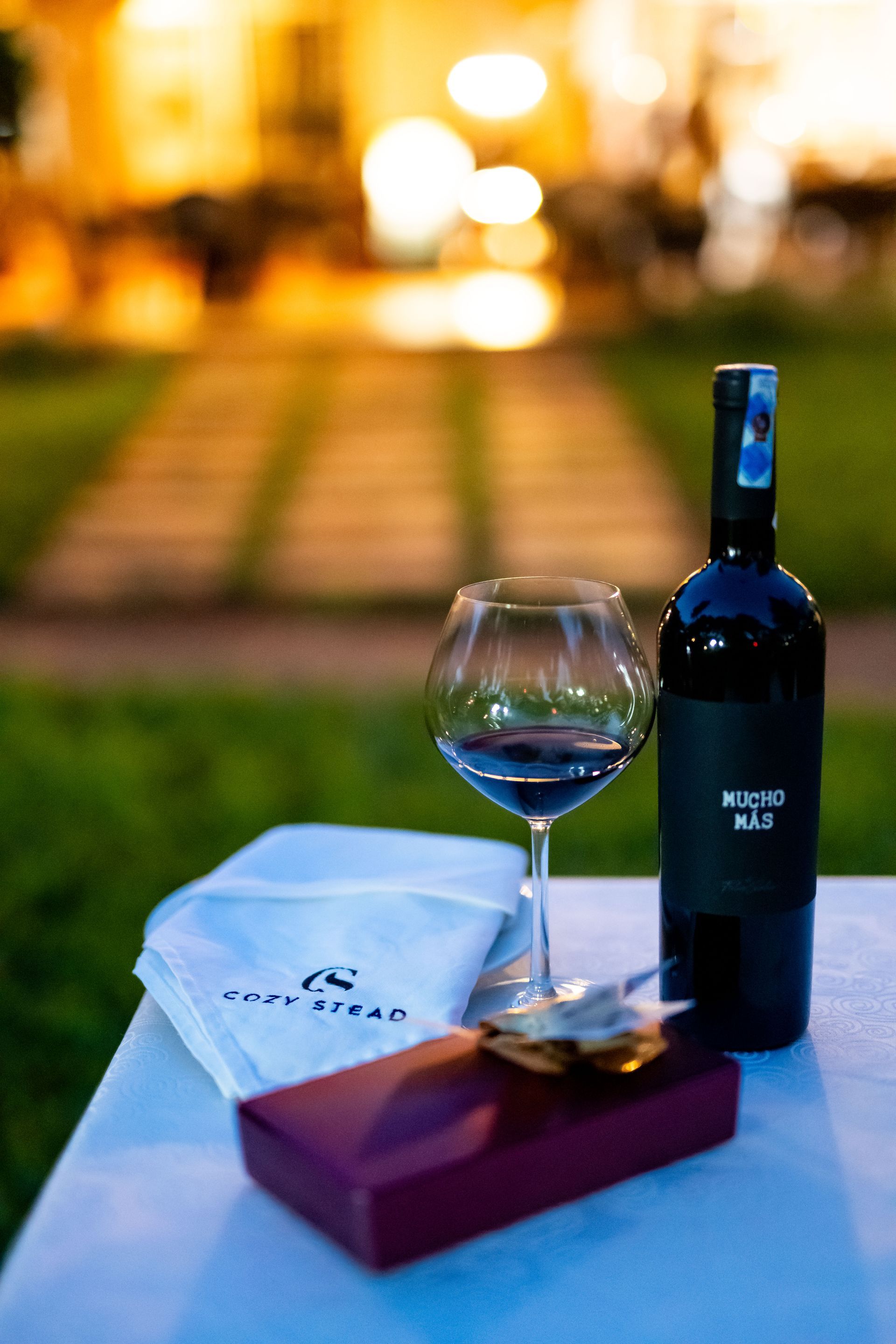 A bottle of red wine and a glass sit on a white table. A gift box and folded cloth are present, set against an outdoor scene.