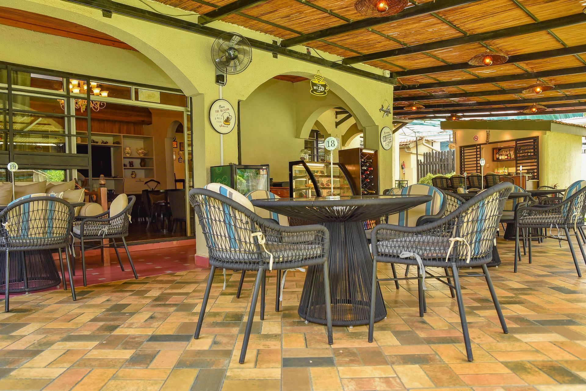 A dining area with wrought iron tables and chairs on a brick-patterned patio, under an arched roof with bamboo ceiling.
