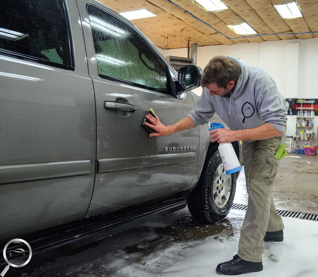 A person in a gray hoodie and tan pants cleans the side of a gray truck in a workshop with soapy water and a sponge.