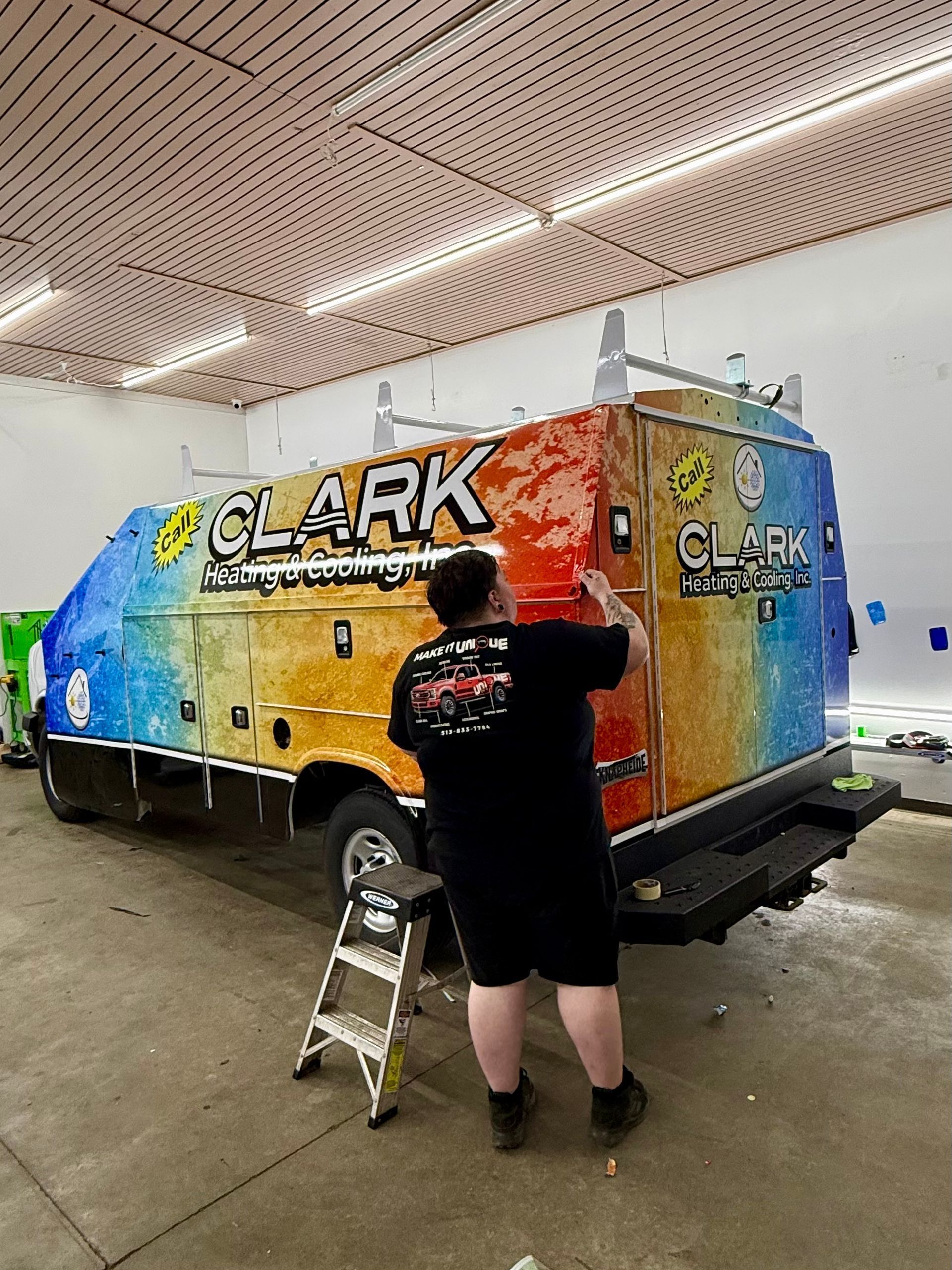 A person in a blue shirt applies a vinyl graphic featuring flames to the side of a white cargo trailer in a workshop.