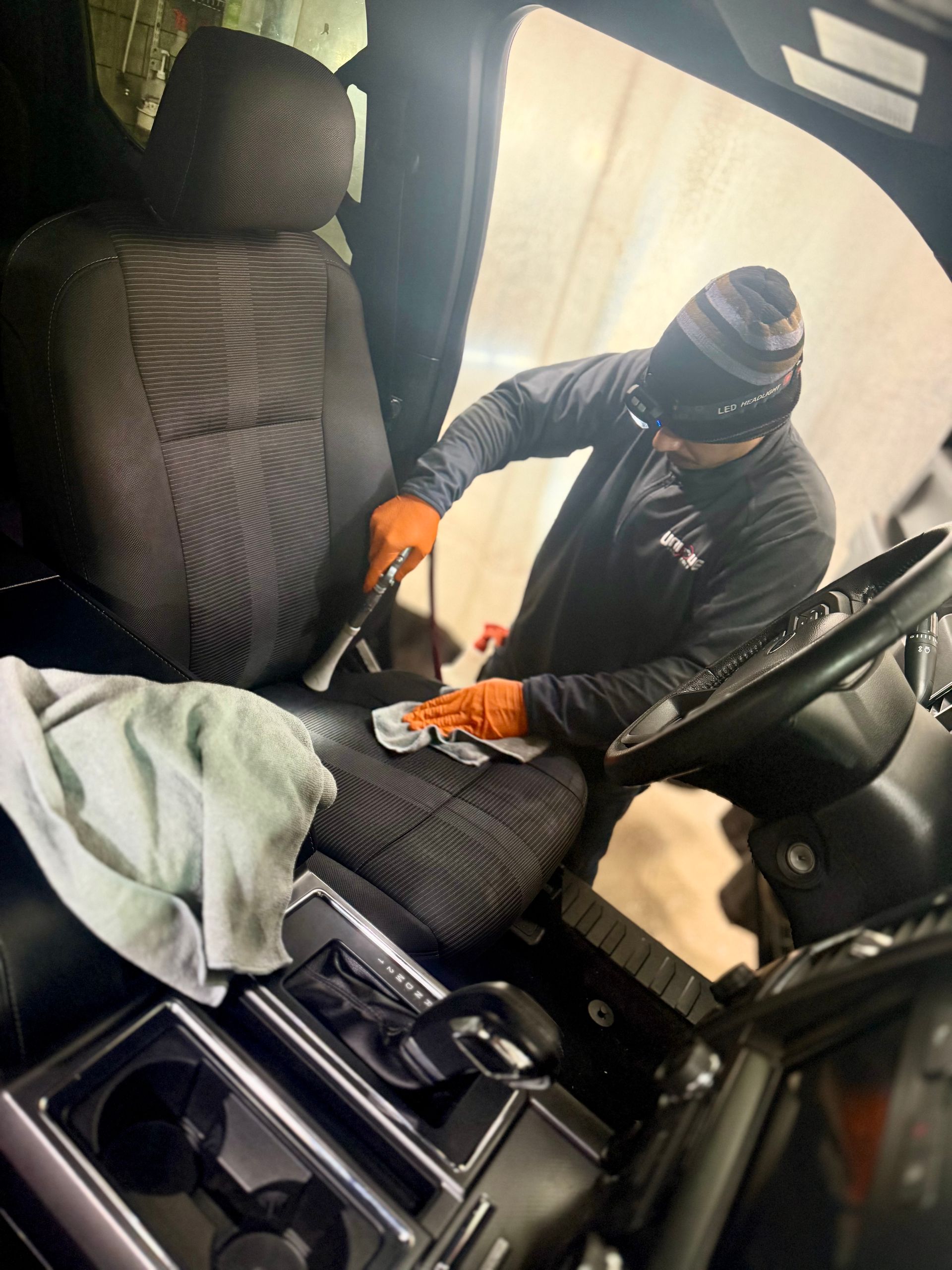 Worker in black beanie and orange gloves cleaning a car seat and interior.