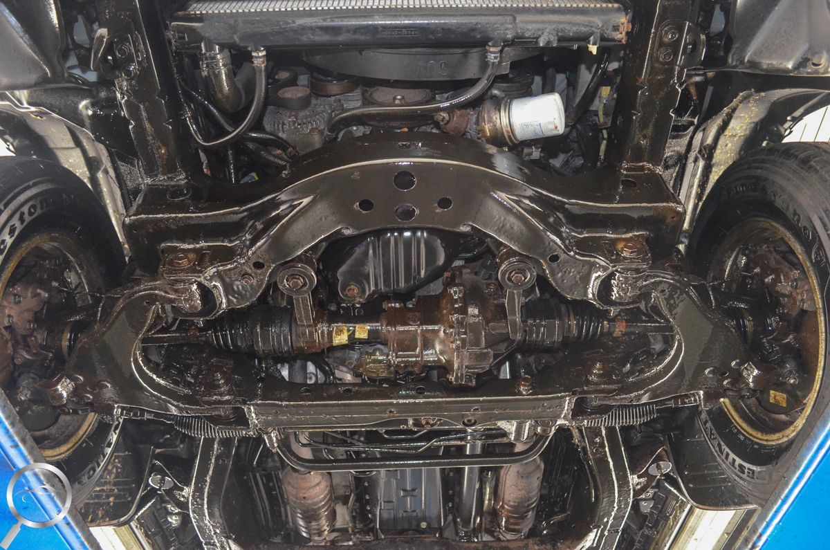 Undercarriage of a car on a lift, showing suspension, drivetrain, and oily mechanical parts