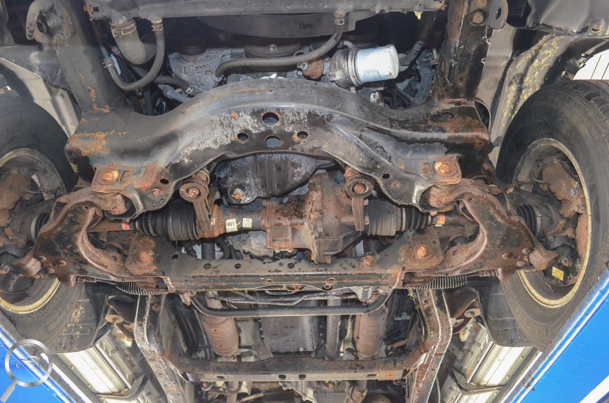 Undercarriage of a car on a lift, showing the rusty front suspension and drivetrain components.