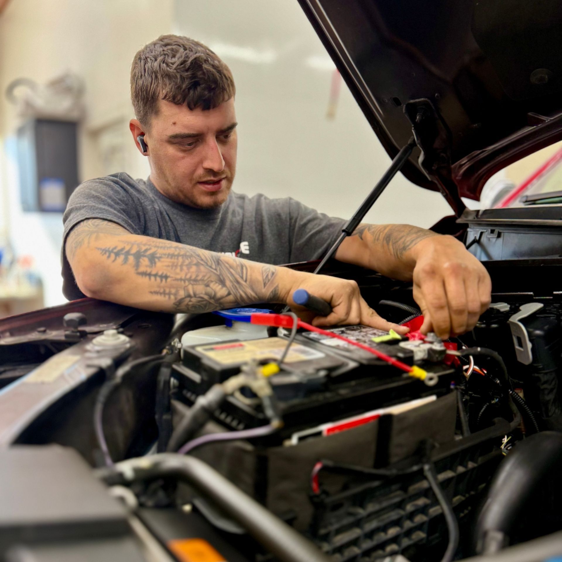 A mechanic with tattoos on their arm is working under the hood of a car, connecting jumper cables to the battery.