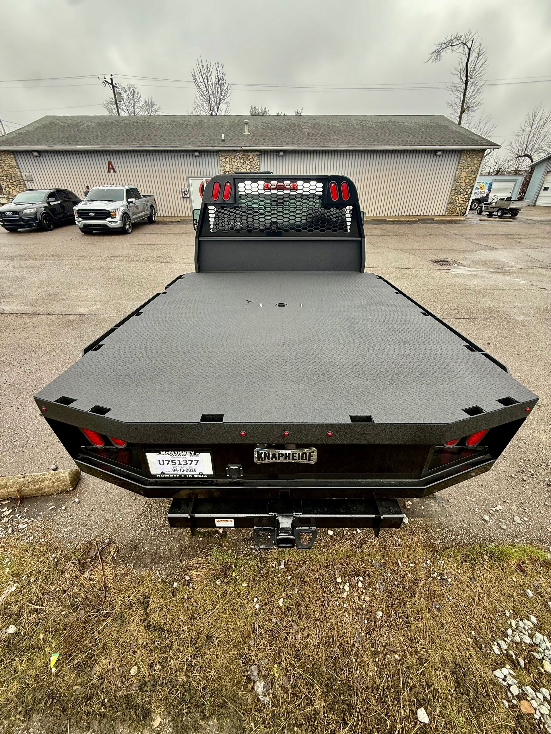 Flatbed truck parked on gravel in front of a beige metal building with cars nearby.