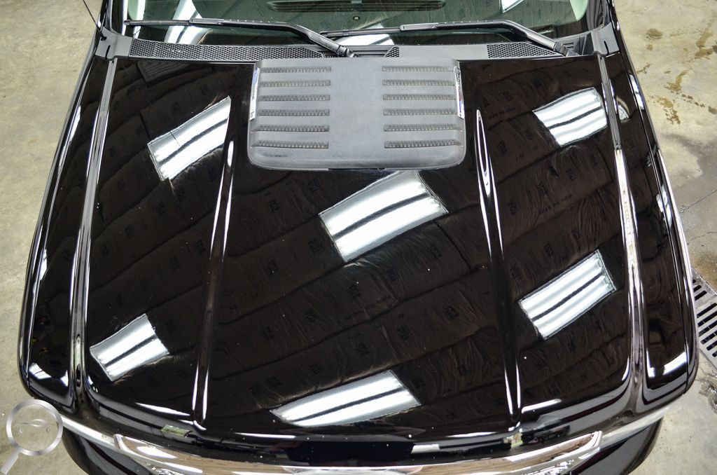 Glossy black car hood with bright ceiling light reflections and a silver vent near the windshield