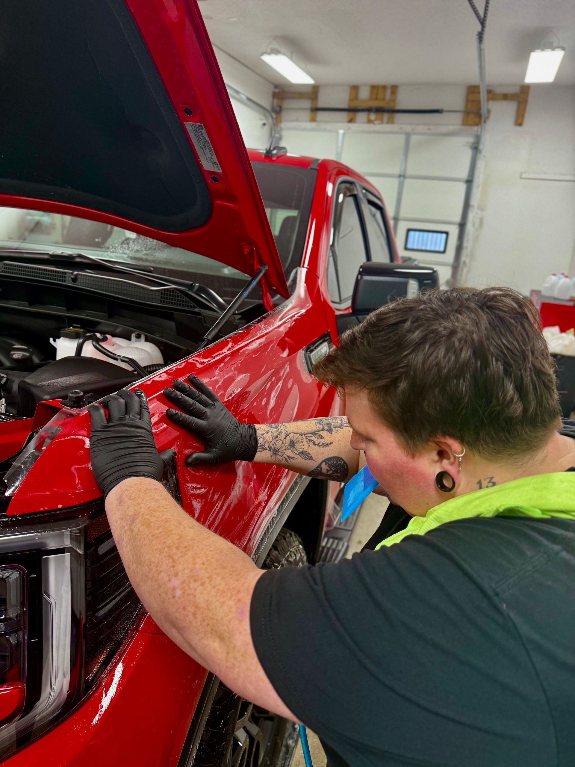 A person with tattoos on their arm wearing black gloves applies a clear protective film to the red fender of a truck.