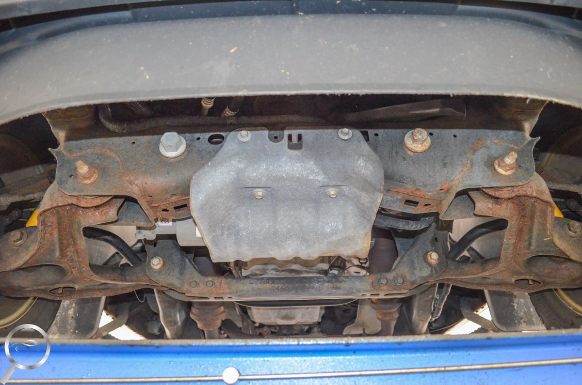 Undercarriage of a vehicle on a lift, showing rusted suspension and a central metal skid plate.