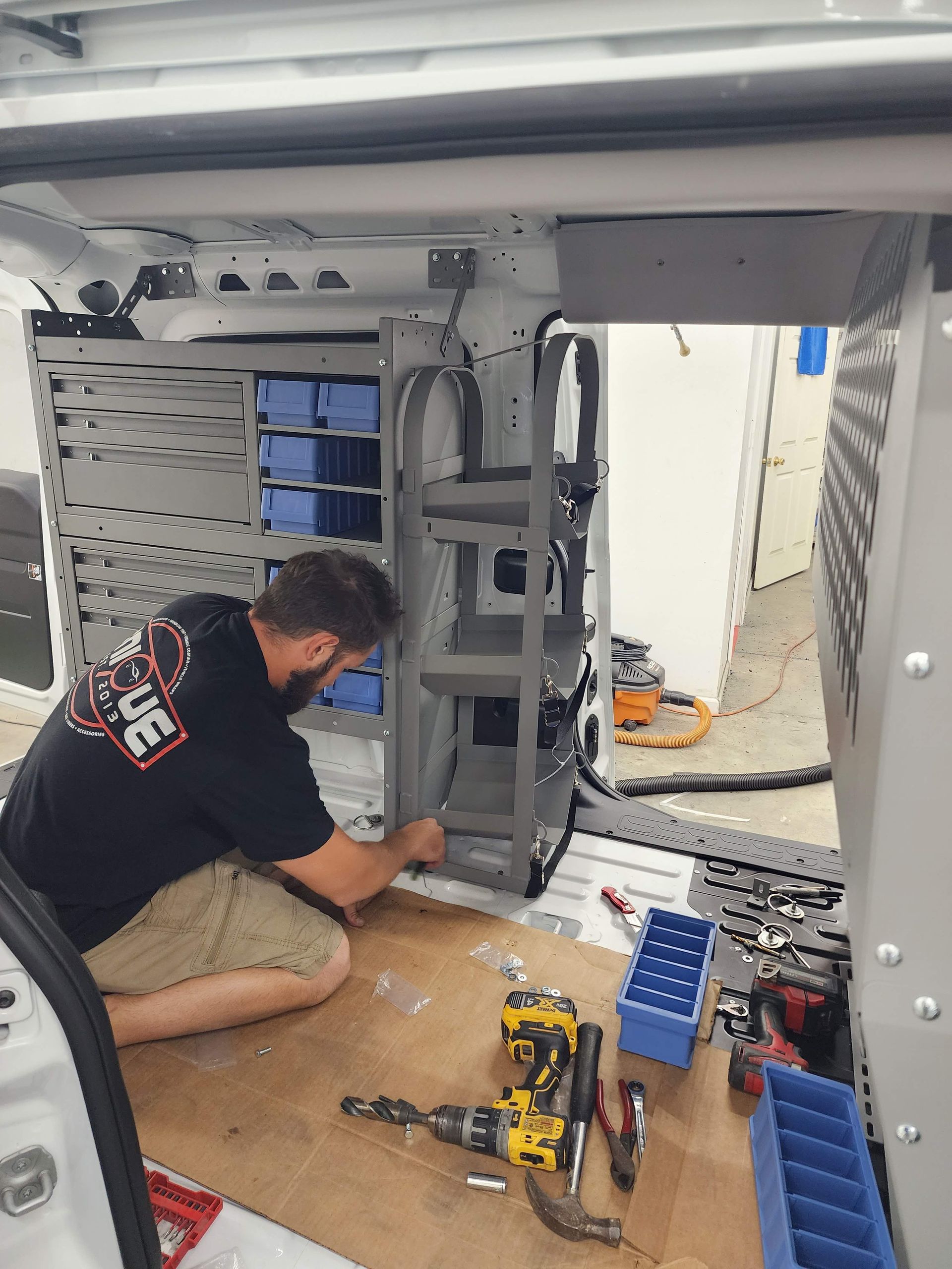 A person kneeling inside a van, installing metal shelving units with a cordless drill and various tools.