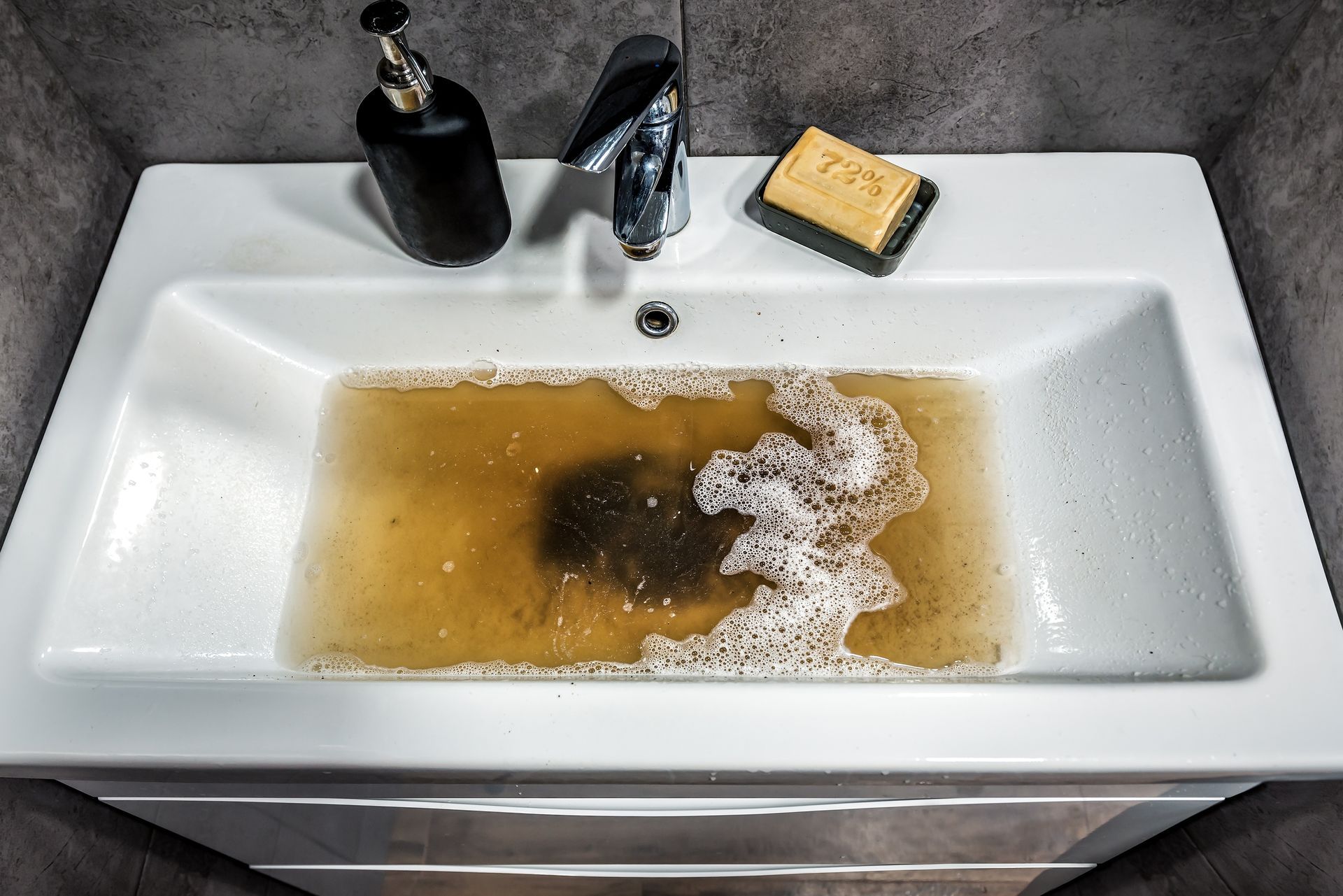White sink filled with brown, dirty water, soap and dispenser on the sides.