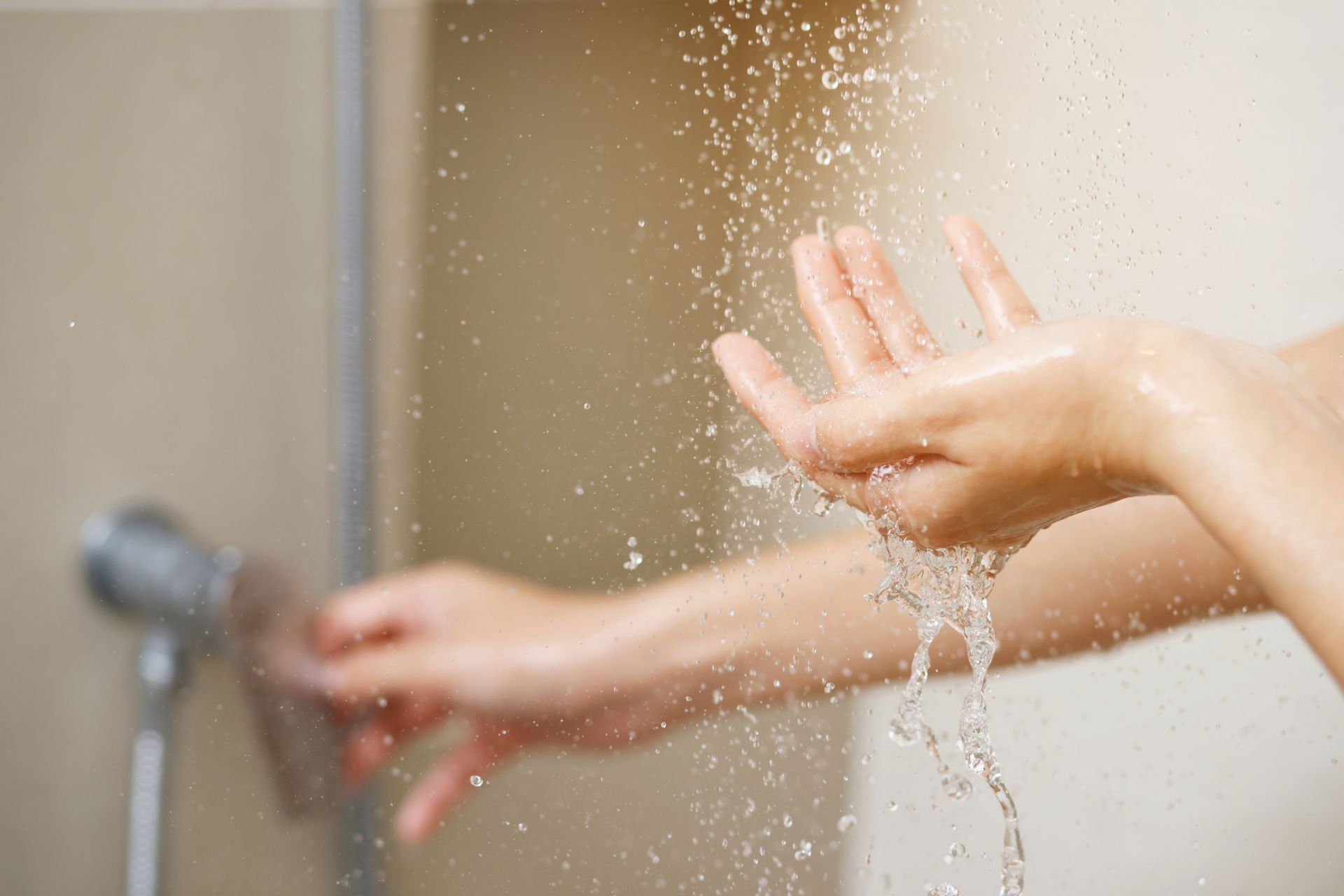 Person's hands under shower spray, testing water temperature; light skin, beige bathroom.