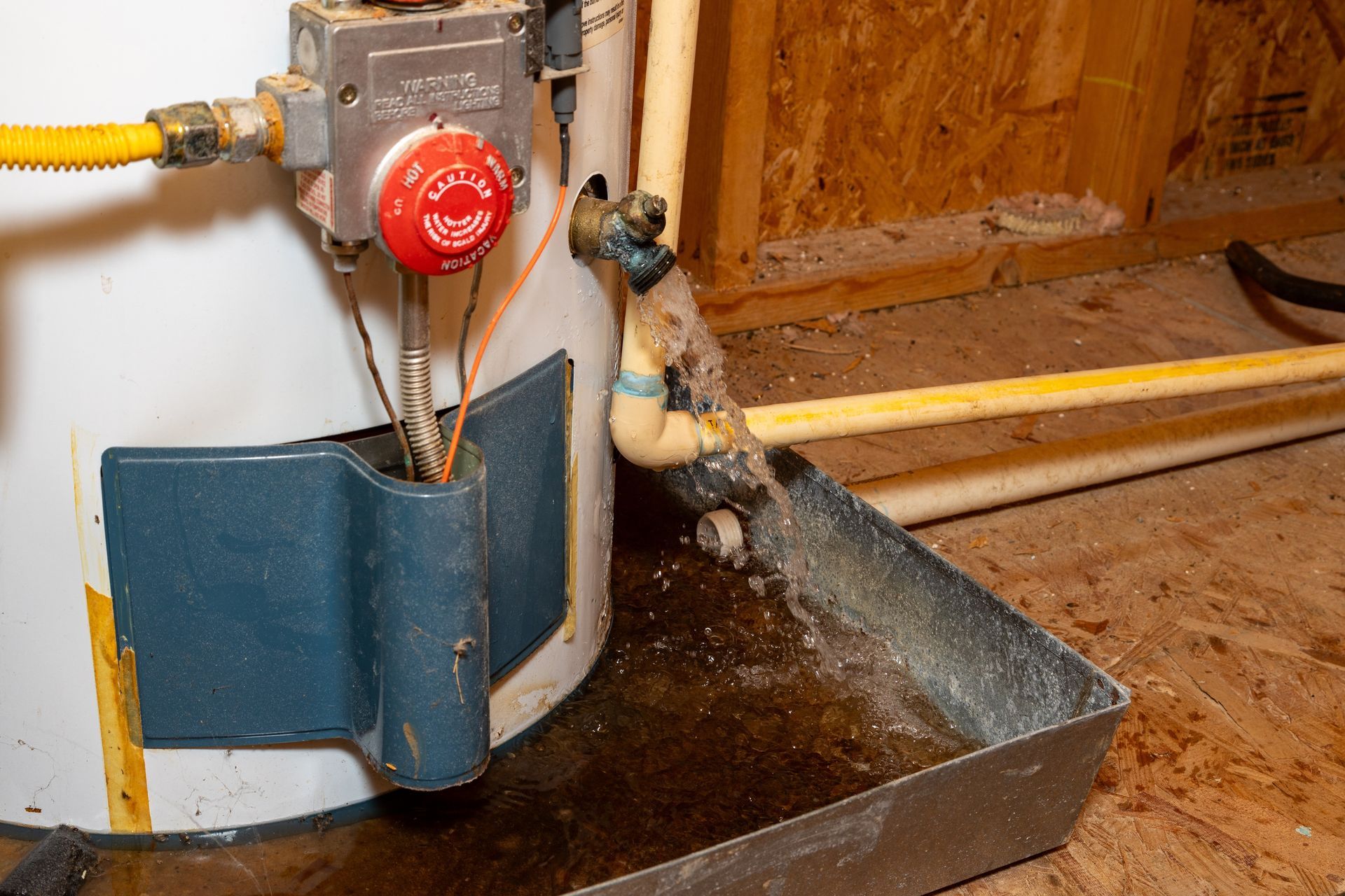 Water heater with a leak into a metal pan in a wooden-walled room. Brown and yellow pipes visible.