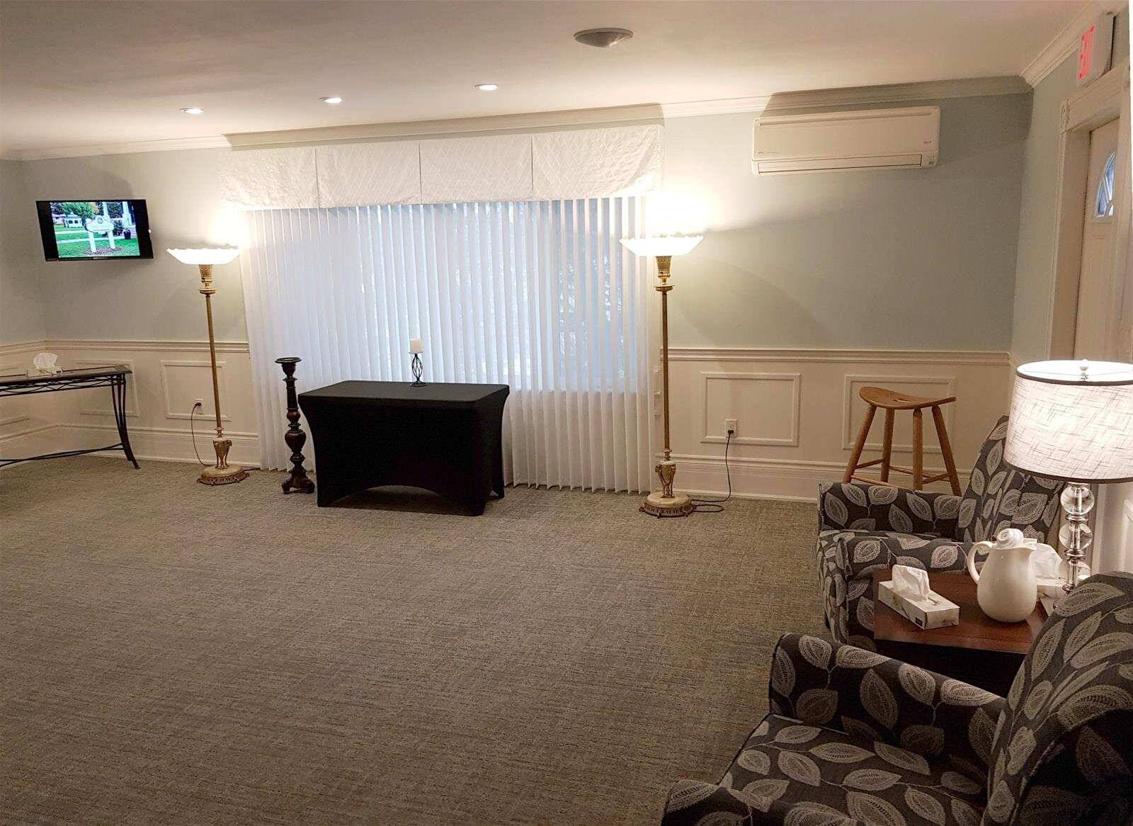 Empty hall with carpet, white curtains, wall lights, a TV, and a black-draped table on a stage.