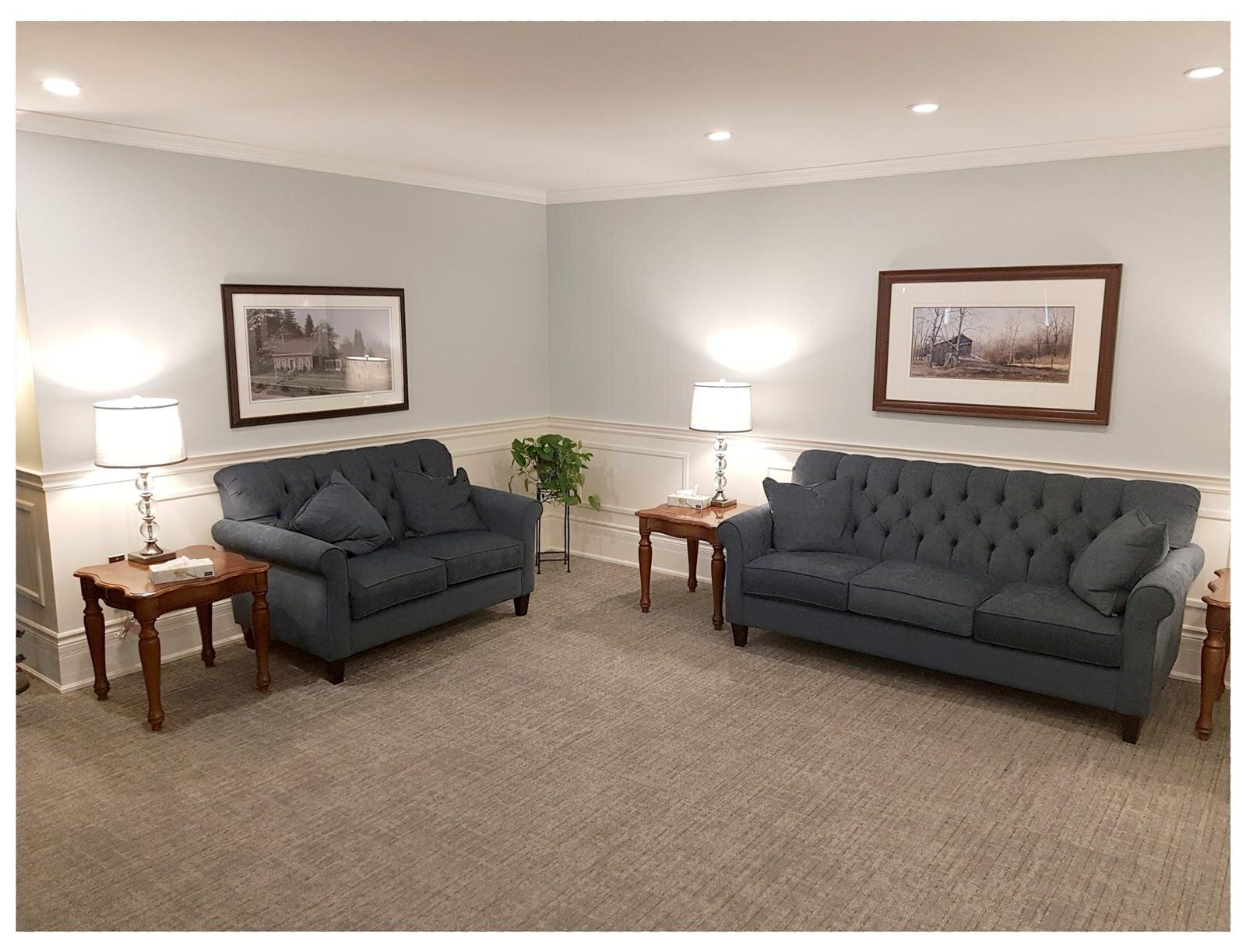 Cozy living room with two dark sofas, side tables, lamps, framed art, and a potted plant on beige carpet.