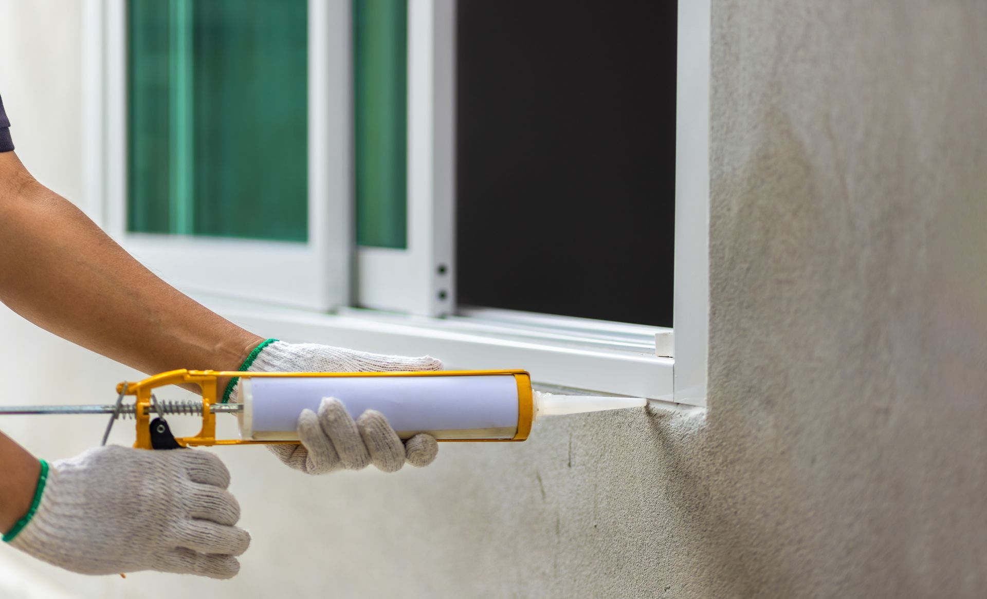 Personne appliquant du mastic sur une fenêtre à l'aide d'un pistolet à calfeutrer ; cadre de fenêtre blanc, mur gris clair.
