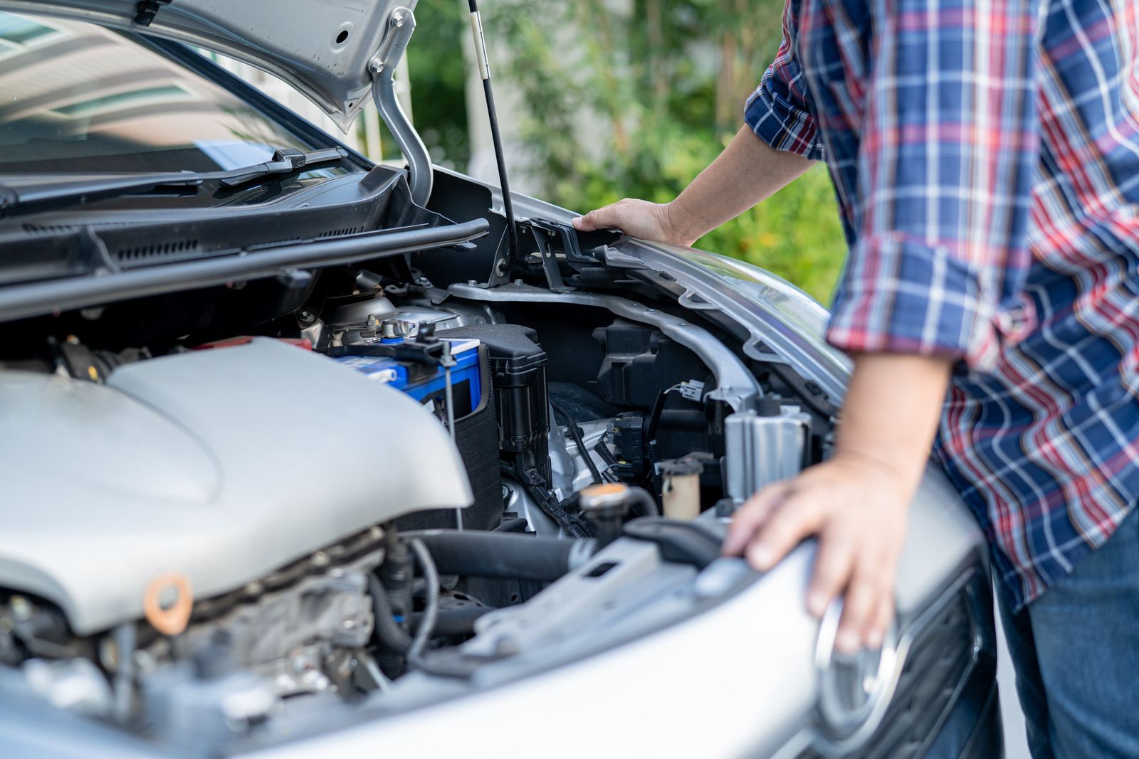 Person inspecting a car engine with the hood open, wearing a plaid shirt.