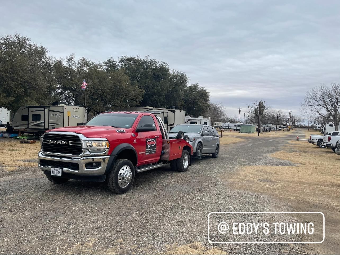 Red tow truck towing a car on a gravel road in a campground with RVs under a cloudy sky.
