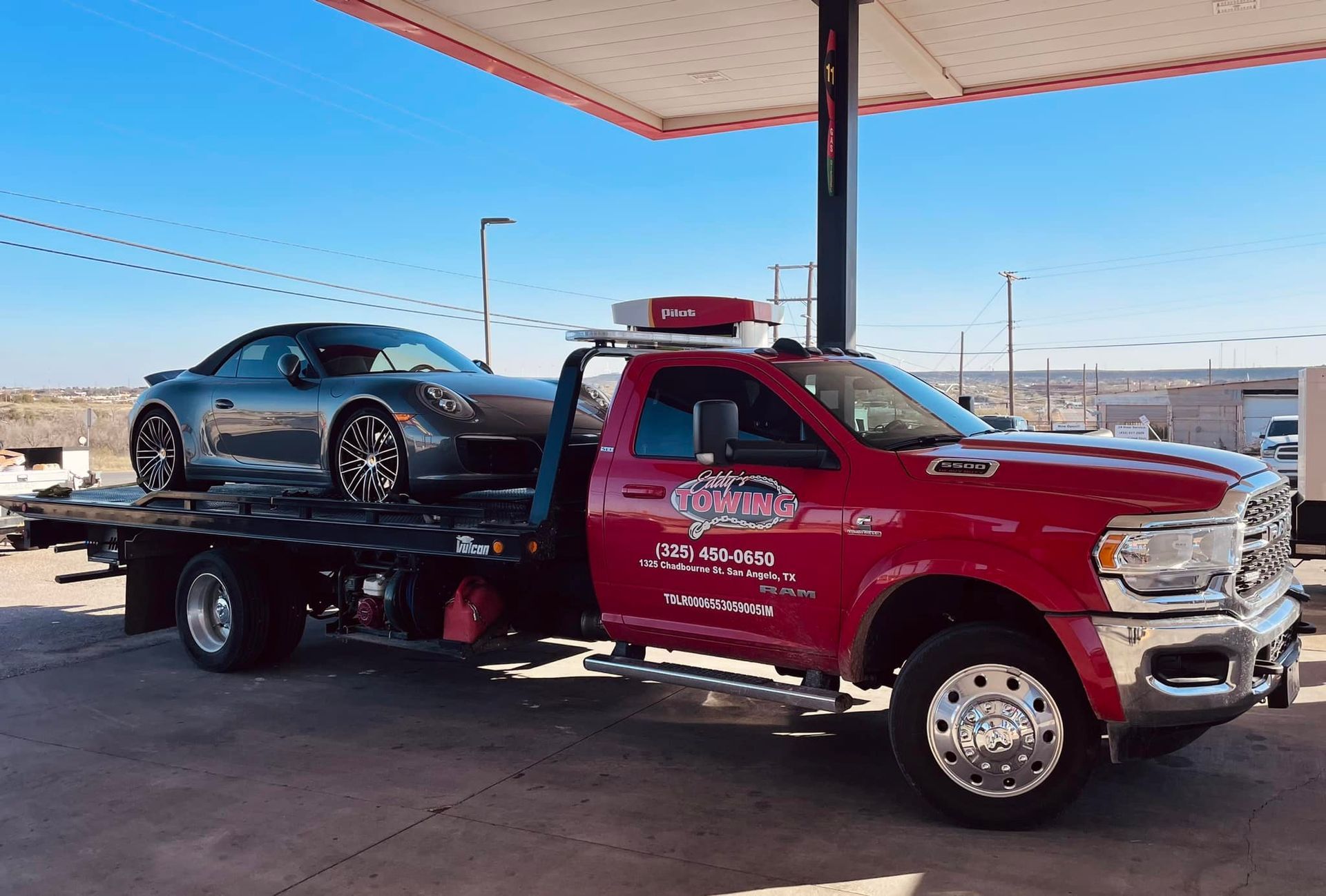 Red tow truck carrying a gray sports car at a gas station under a canopy.