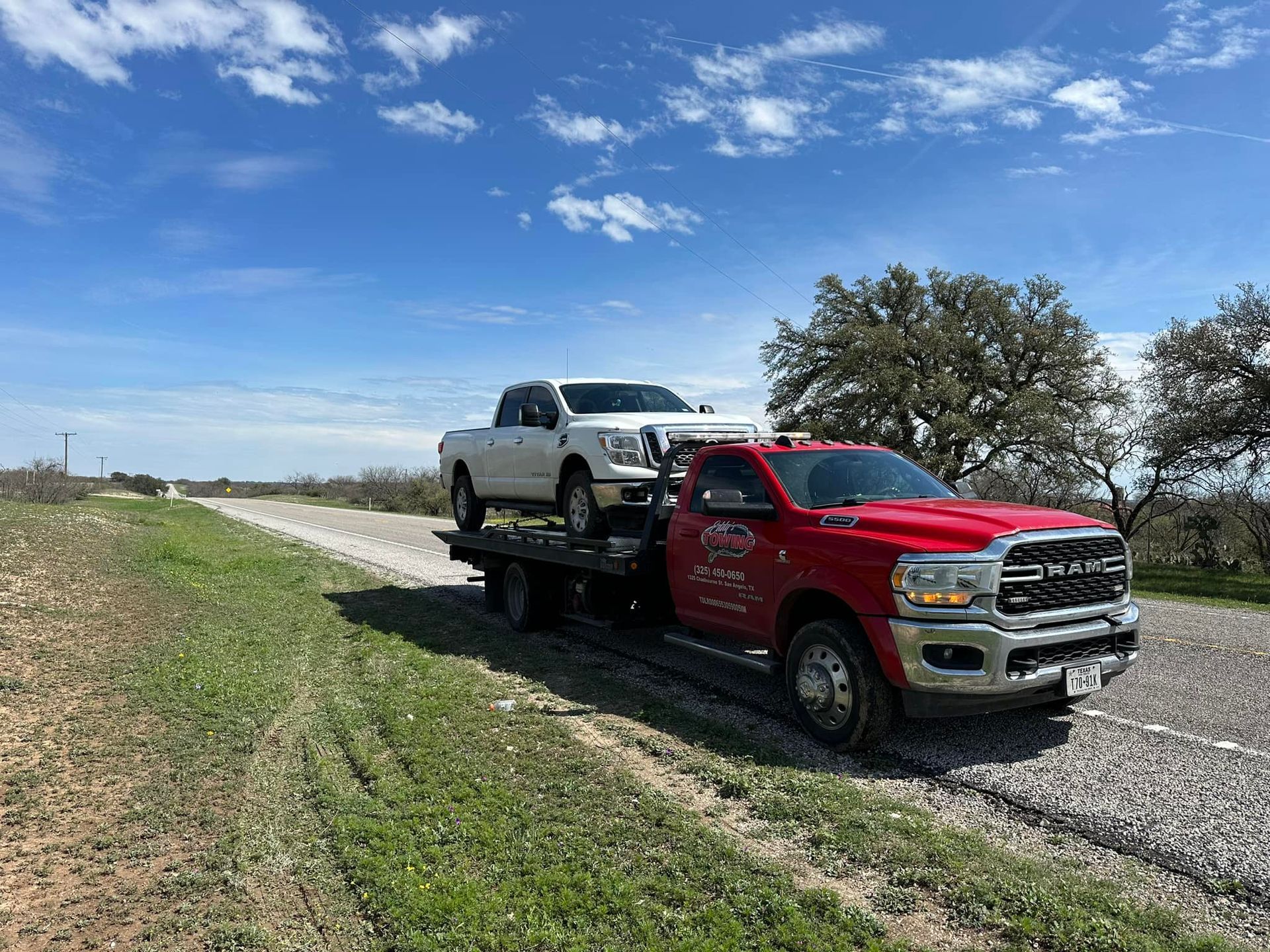 A white pickup truck being towed by a red tow truck on a gravel road, under a blue sky.