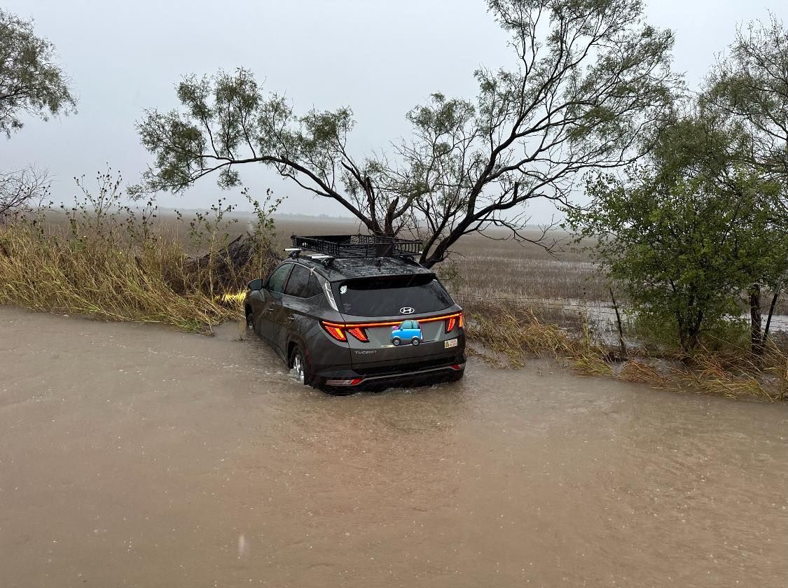 Car partially submerged in floodwater on a road, gray vehicle, overcast sky, brown water, trees and tall grass.
