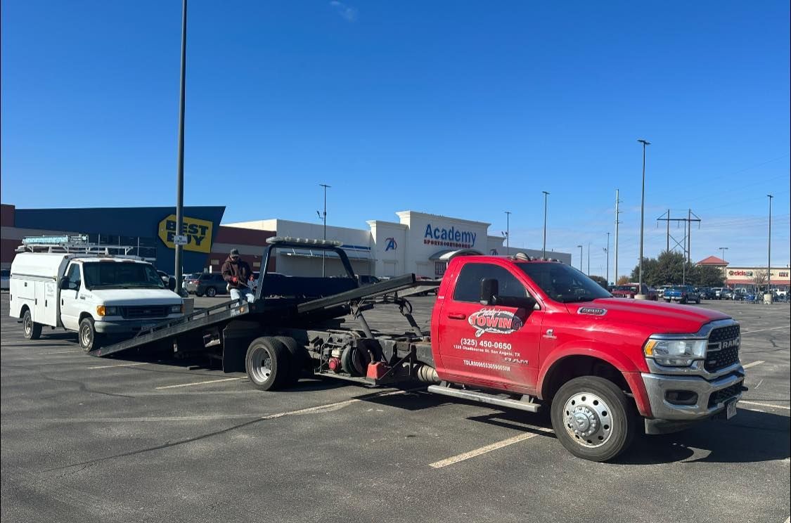 Red tow truck towing a white work van in a parking lot. Blue sky, buildings in background.