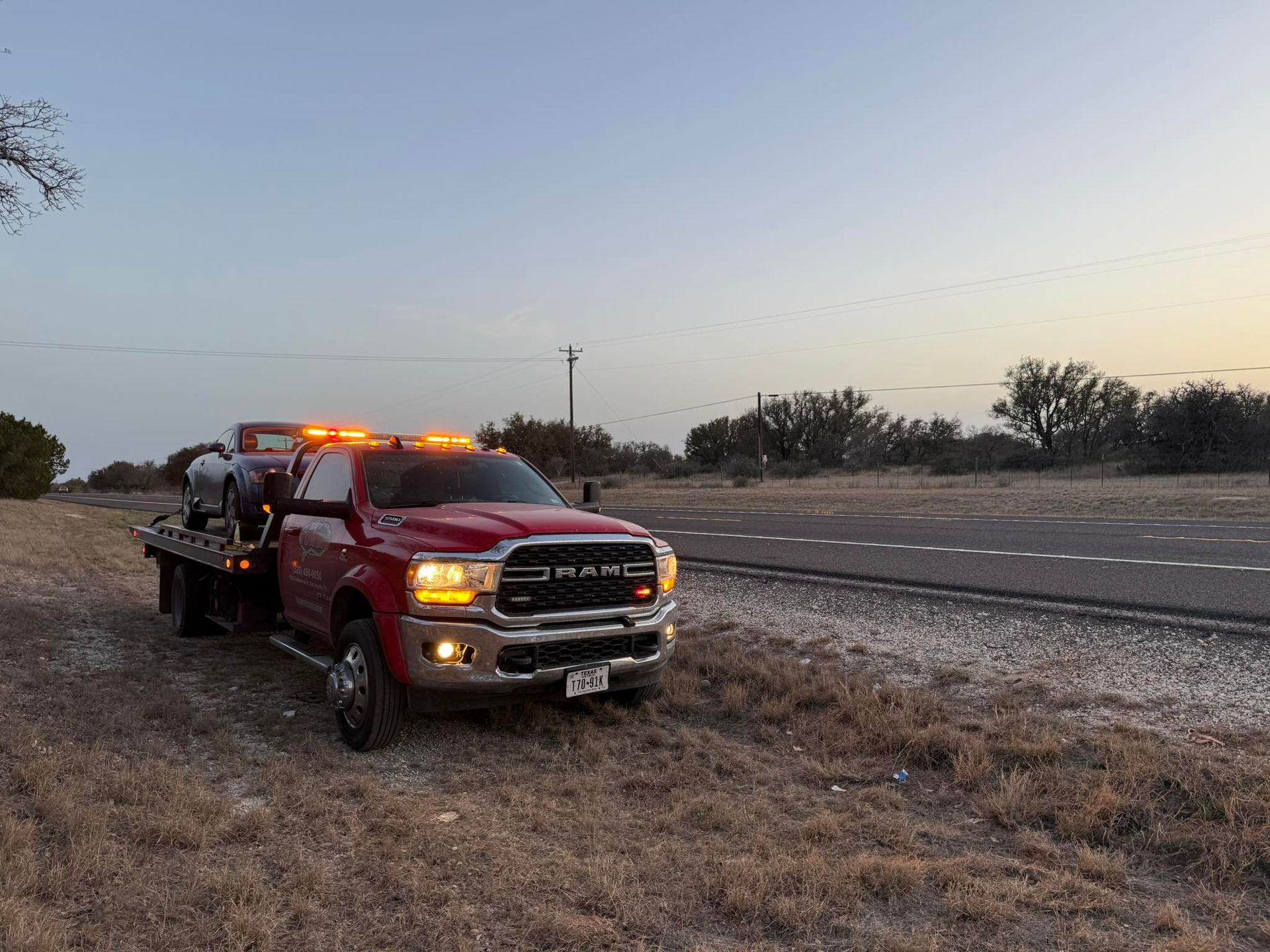 Red tow truck with a car on the flatbed, parked next to a road, at dusk.