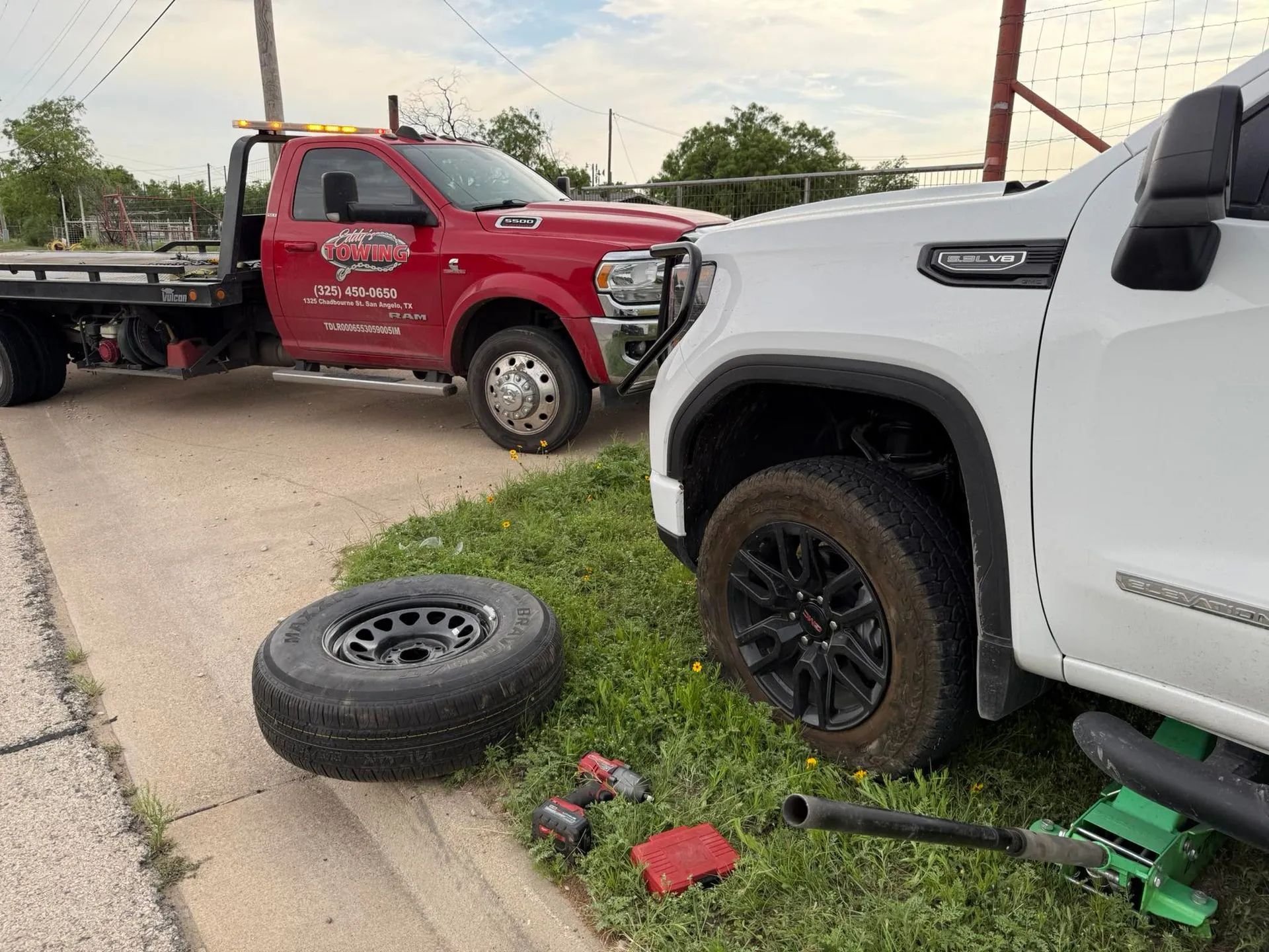 White truck with a flat front tire beside tools, loose wheel, and a red tow truck on a roadside