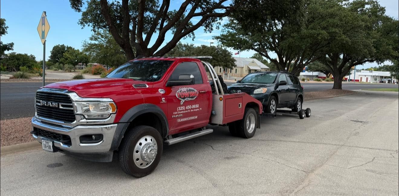 A red tow truck towing a black SUV on an asphalt parking area, trees in background.