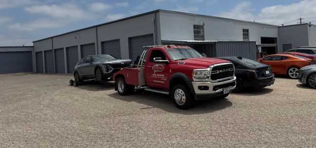 Red tow truck towing a damaged gray car in front of a gray building and other cars.