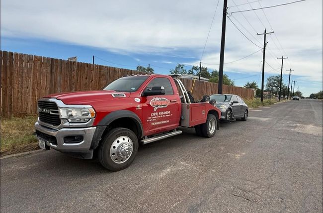 Red tow truck towing a gray car on a city street; wooden fence and power lines in the background.